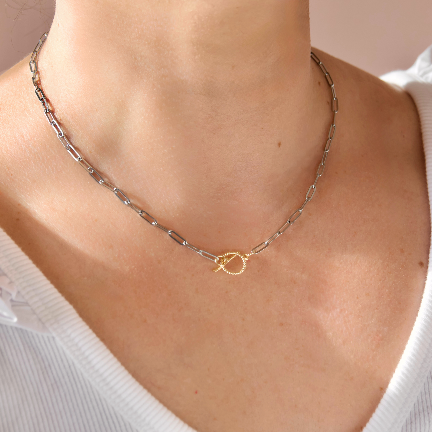 A close-up of a person's neck and upper chest wearing a delicate silver paperclip chain necklace with a small gold infinity symbol charm at the center.