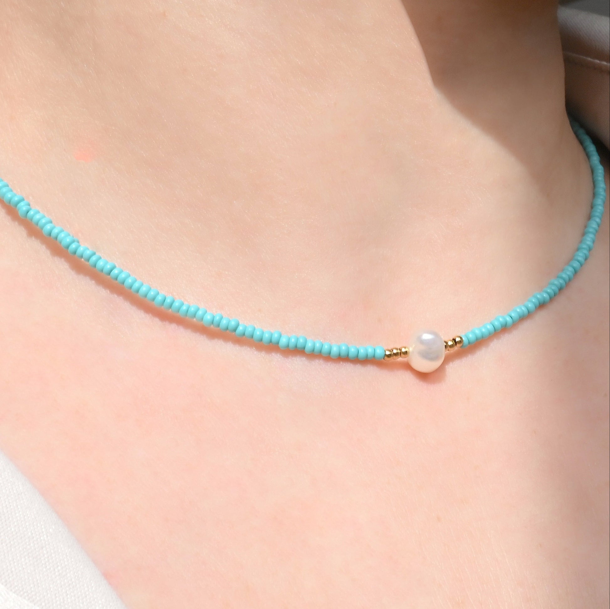 Angled close up view of turquoise bead necklace with a pearl pendant on a person's neck under direct sunlight