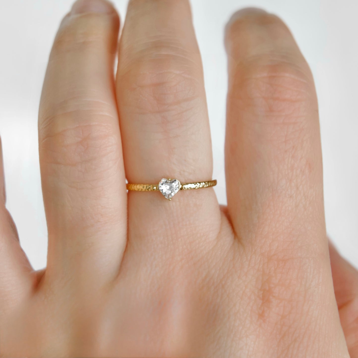 A delicate gold ring with a small heart-shaped clear gemstone, shown worn on a person's finger against a light background.
