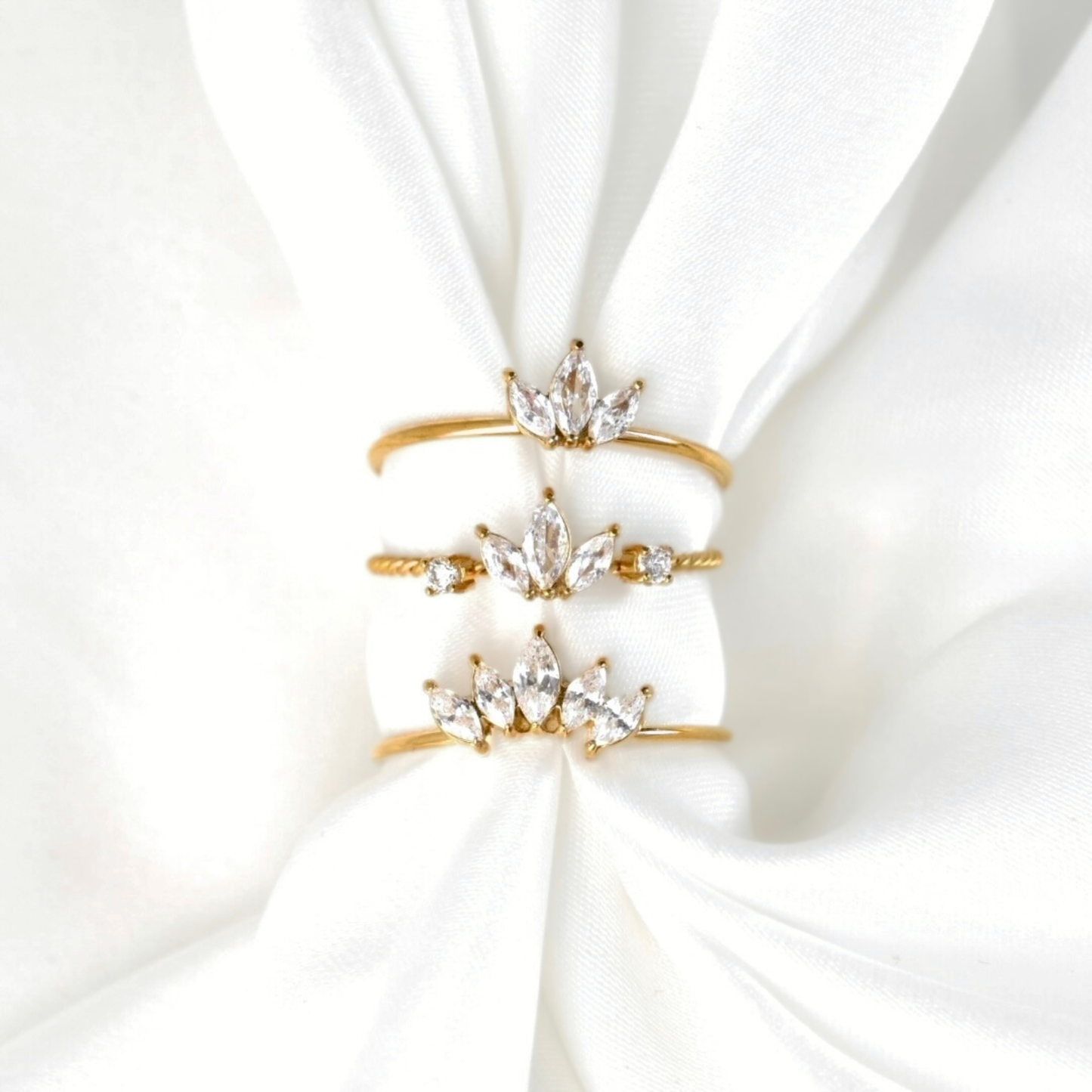 Three gold crown-shaped rings with clear crystal accents, stacked on a white satin fabric background.