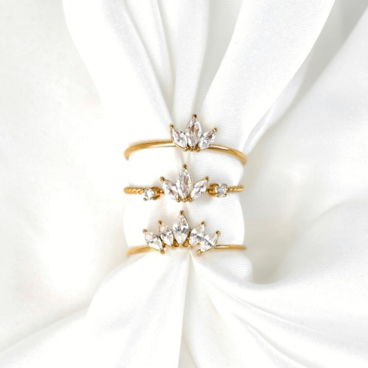 Three gold crown-shaped rings with clear crystal accents, stacked on a white satin fabric background.