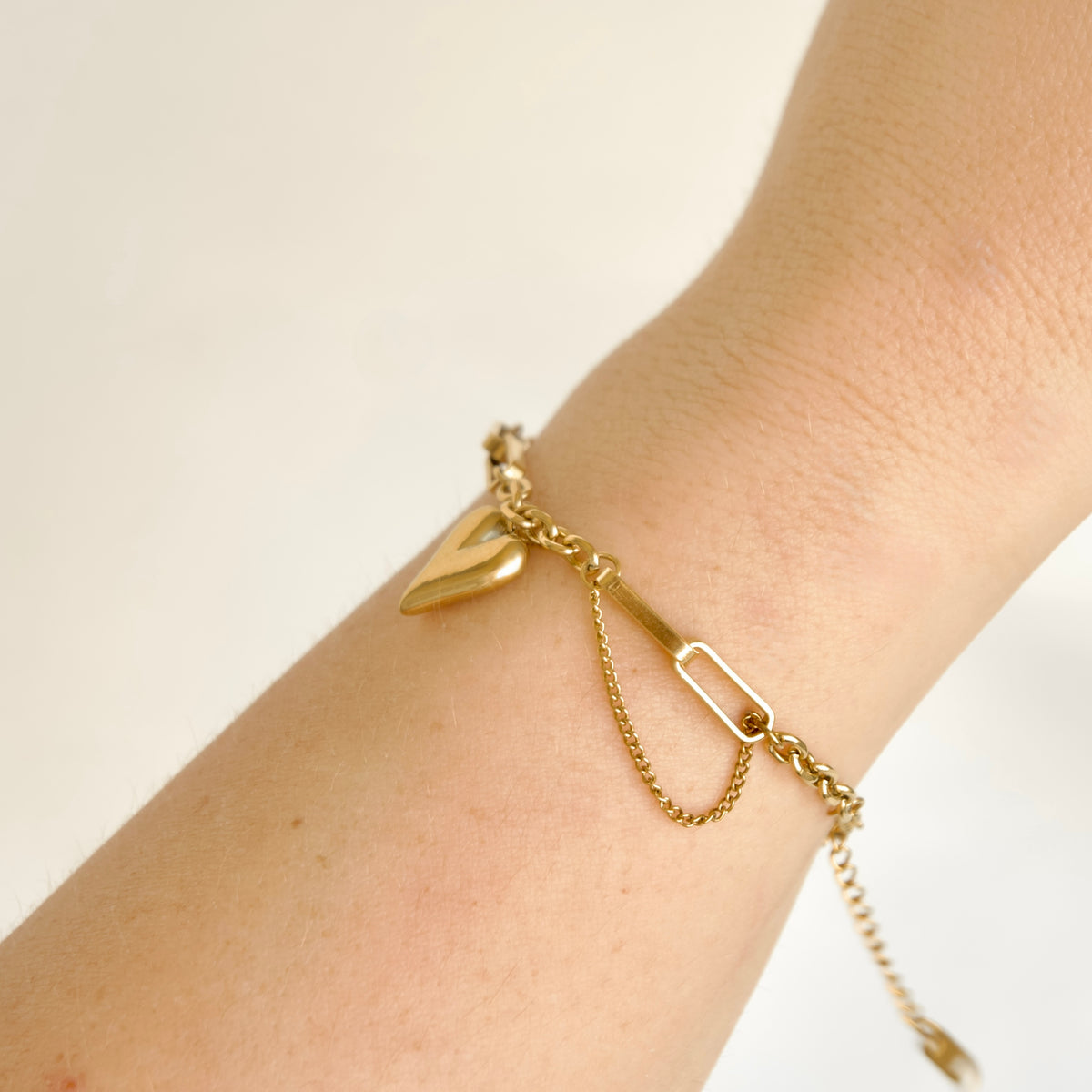 Side profile of a delicate gold chain bracelet with a small gold heart charm and a rectangular link accent, shown worn on a person's wrist against a soft off-white background.