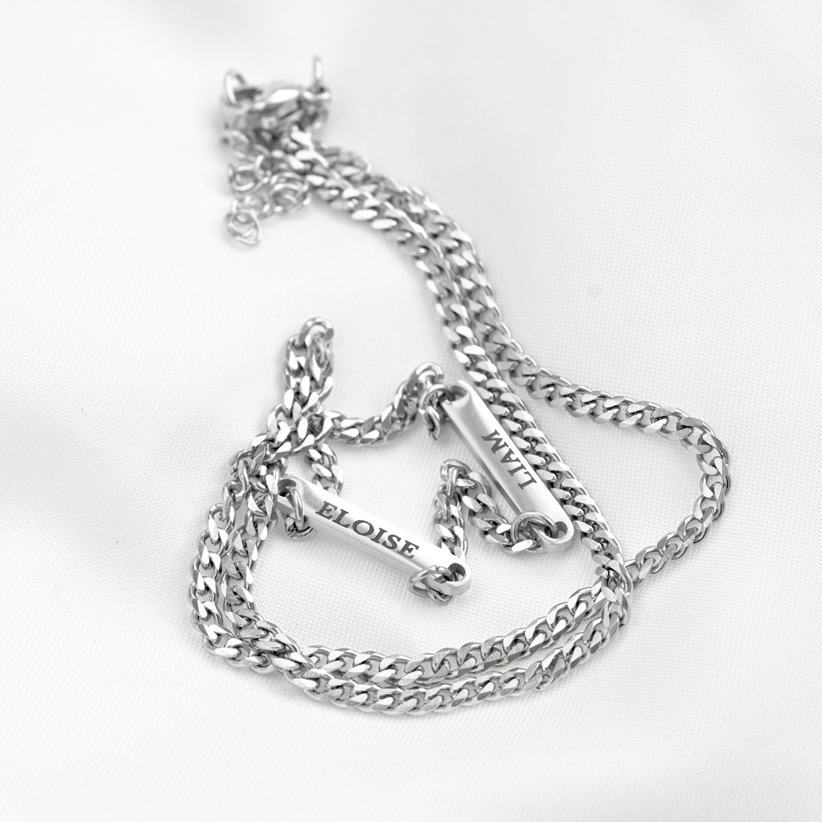 A silver-toned cuban link chain necklace and two nameplates engraved with 'LIAM' and 'ELOISE', displayed on a white fabric background.