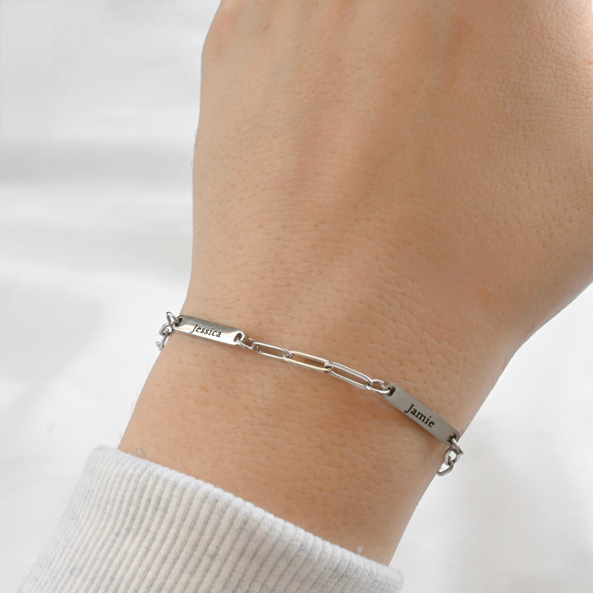 A close-up of a person's wrist wearing a silver  paperclip chain bracelet with engraved names, 'Jessica' and 'Jamie', on a white ribbed sleeve against a soft white background.