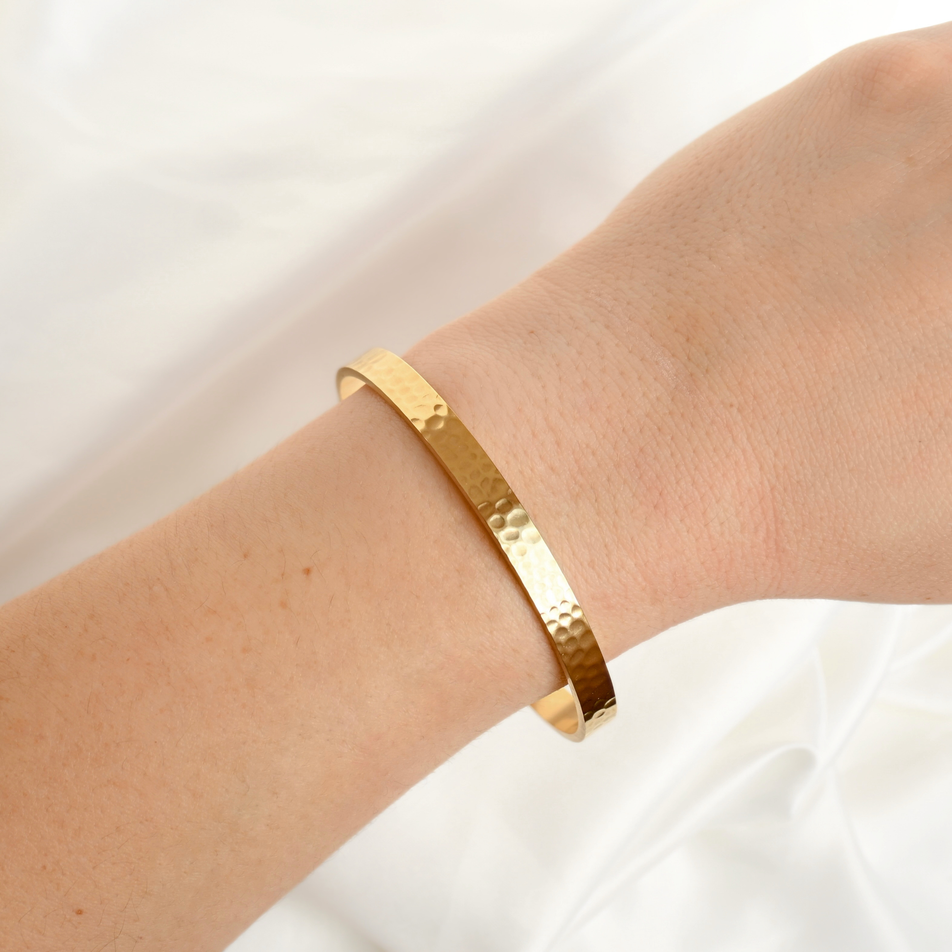 A gold hammered metal cuff bracelet worn on a person's wrist, set against a soft white fabric background.