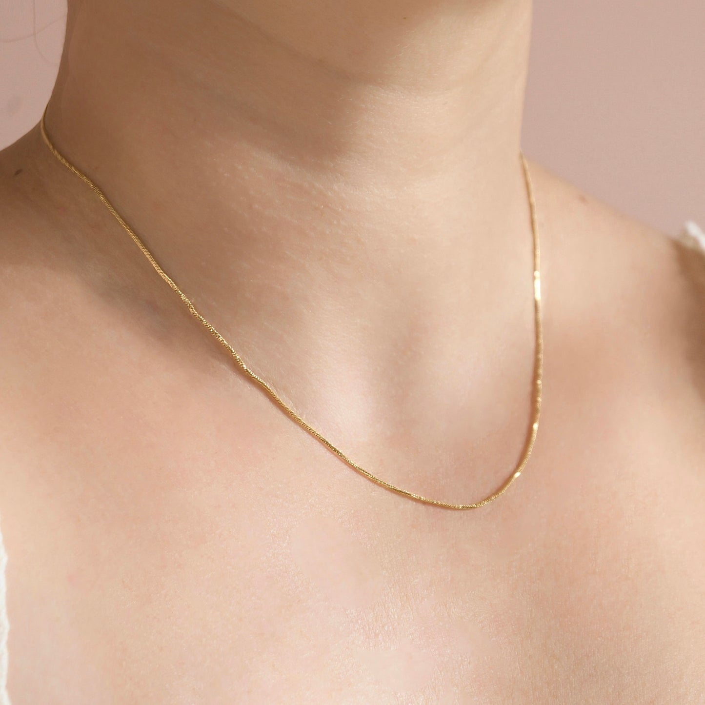A delicate gold chain necklace featuring a thin twisted snake chain worn on a model's neck, shown in a close-up shot against a soft pink background.