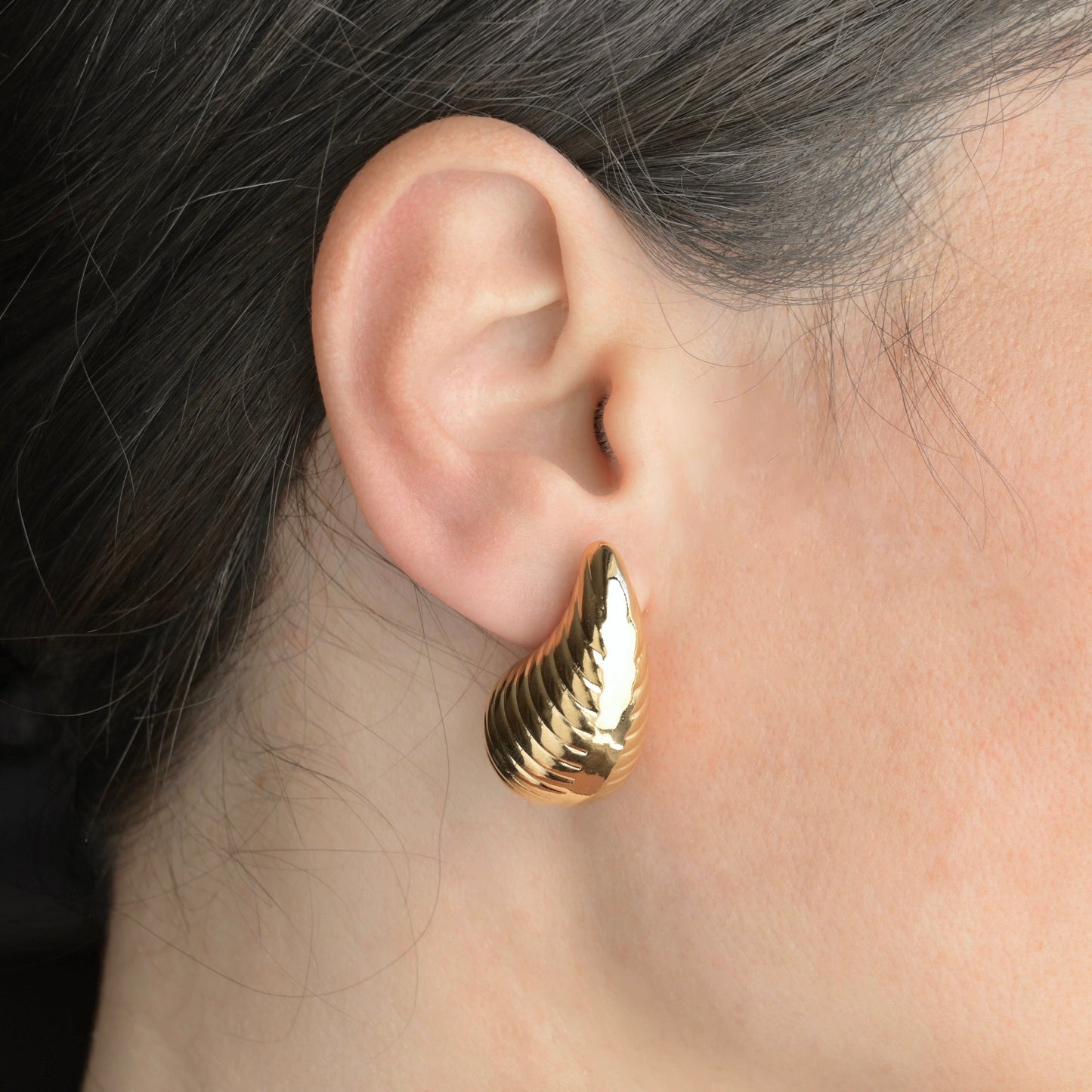 Chunky gold drop earring with ribbed texture worn by a person with dark hair.