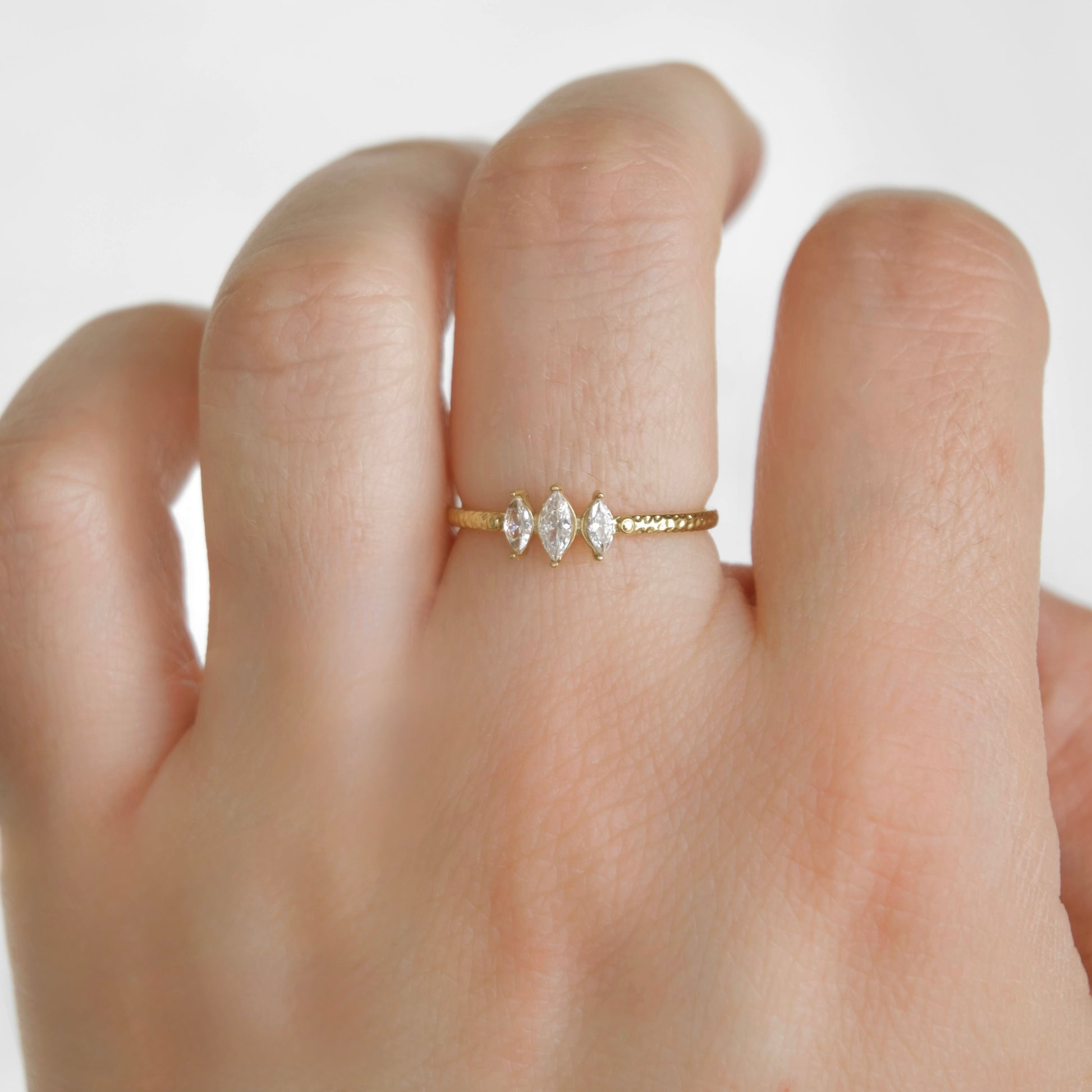 A close-up of a hand wearing a delicate gold ring with three marquise-cut clear gemstones in the center, set against a plain light background.