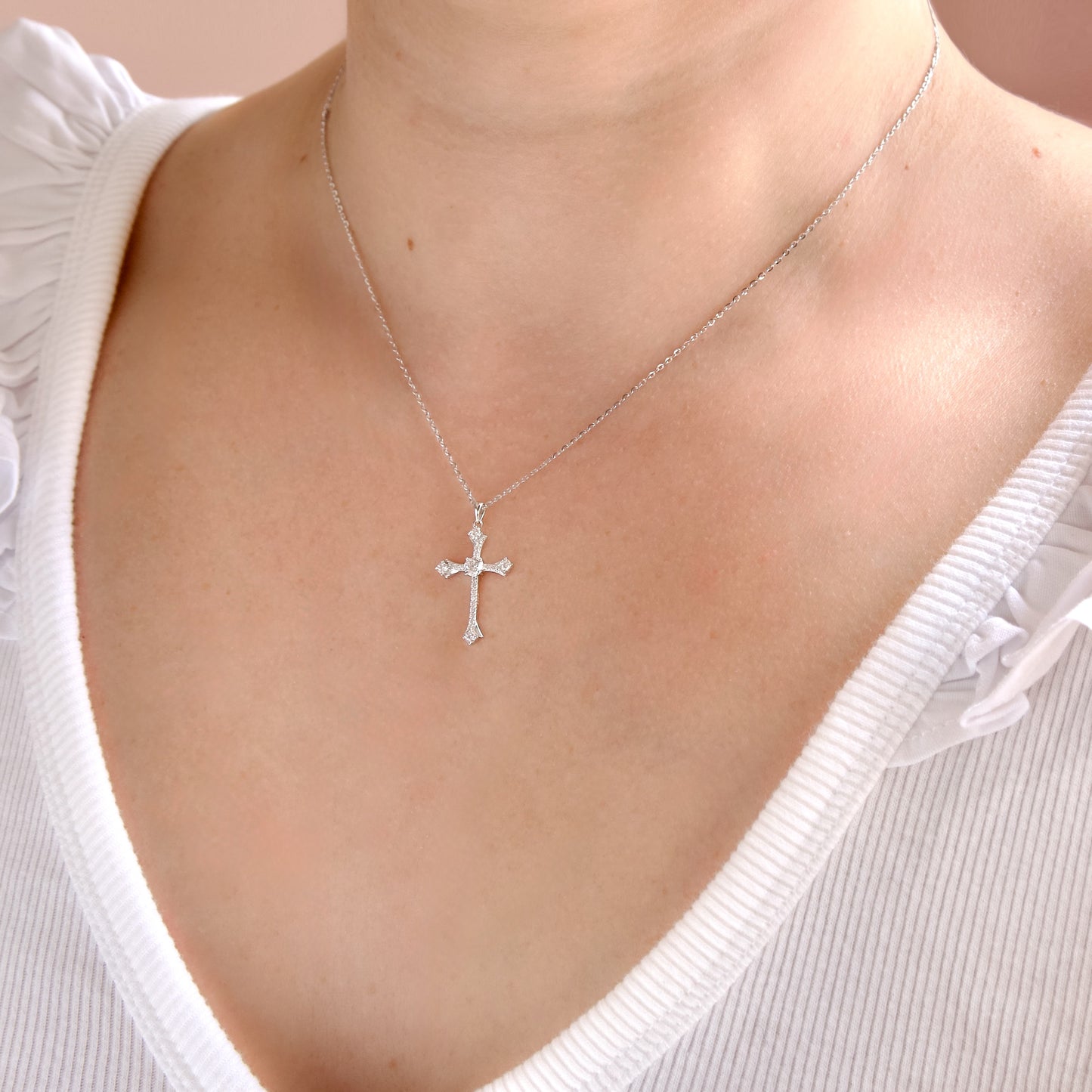 A silver cross pendant necklace with small clear stones, worn on a model's neck over a white ribbed top.