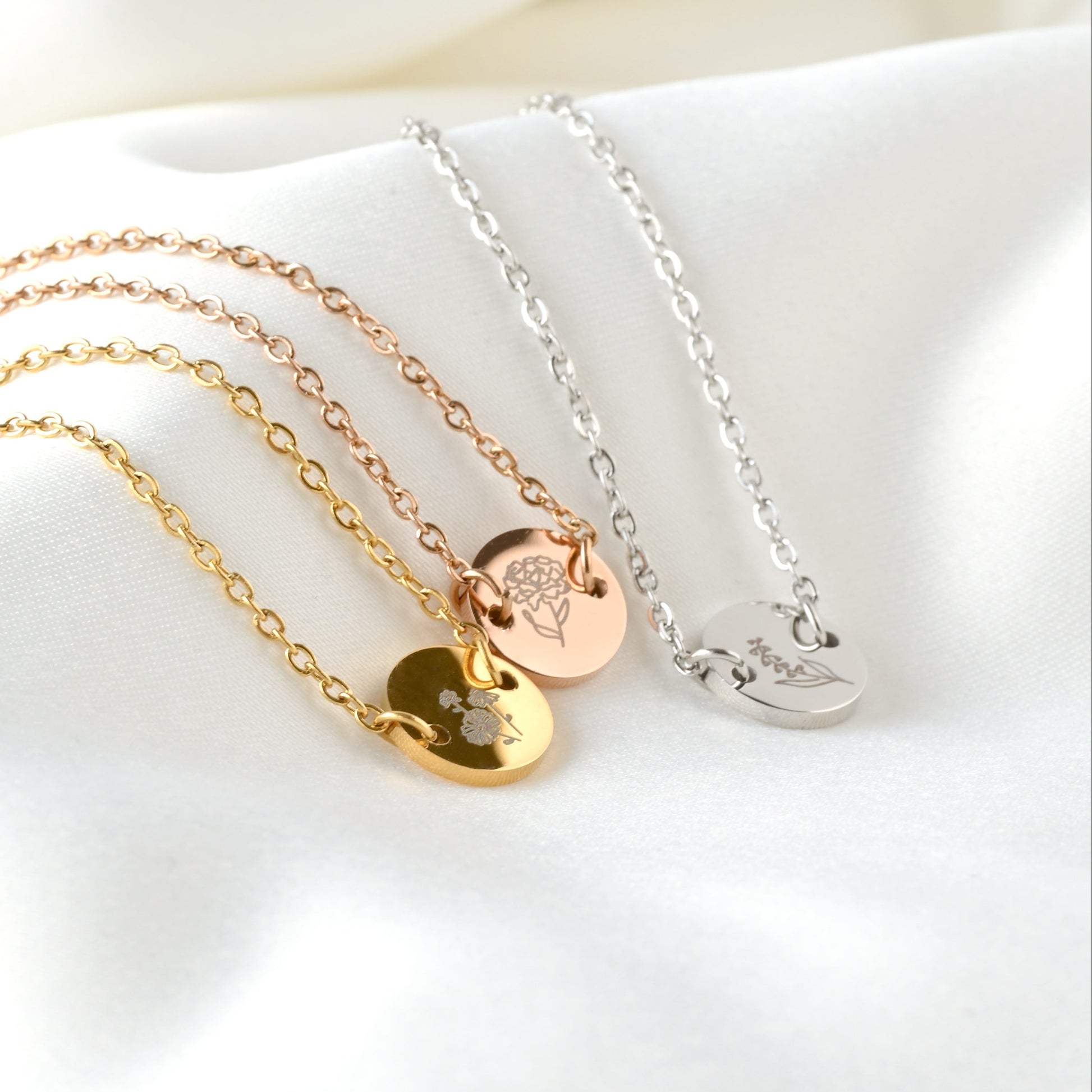 Three bracelets with round birth flower charms in gold, rose gold and silver on a white surface