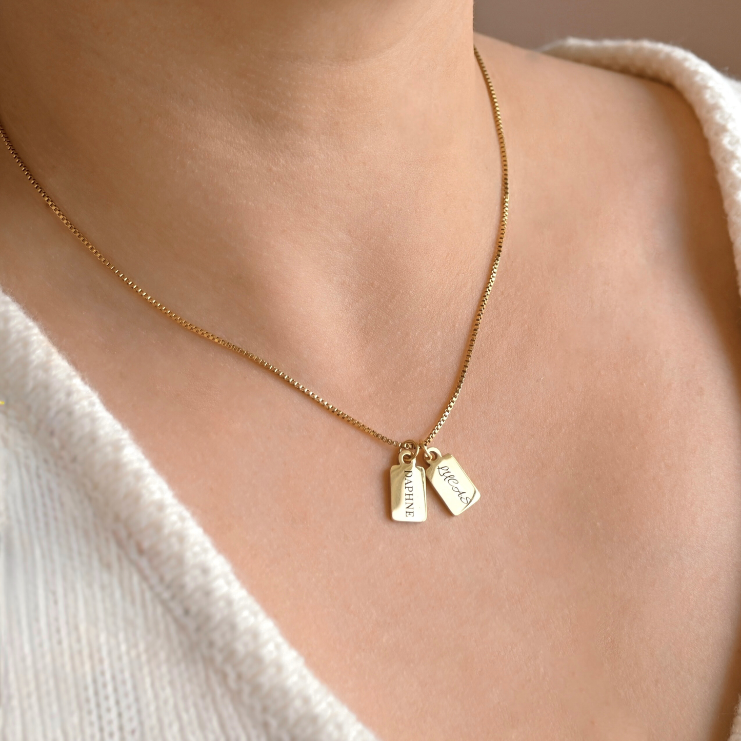 A close-up of a person wearing a delicate gold box chain necklace with two small rectangular engraved gold pendants, one reading 'LUCAS' and the other 'DAPHNE'.