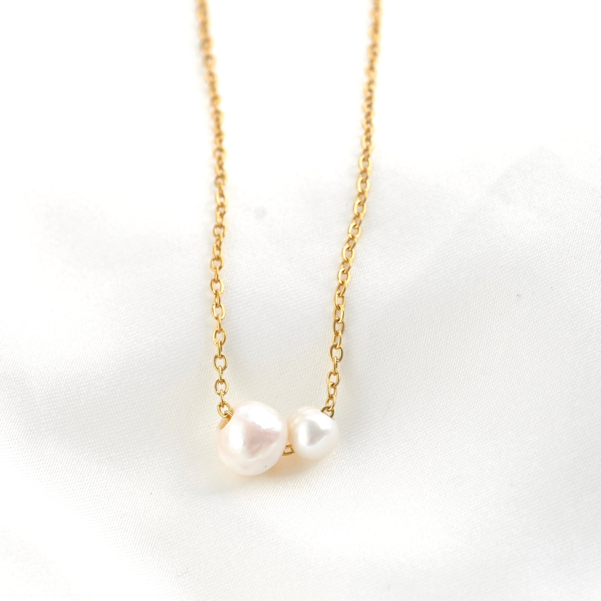 A delicate gold chain necklace featuring two small, irregular white baroque pearls, displayed on a soft white fabric background.