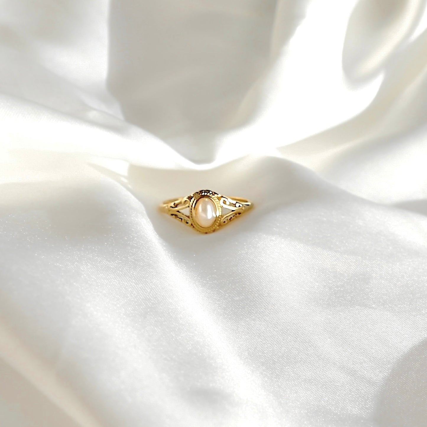 A gold ring with a central white pearl and delicate filigree details, resting on draped white satin fabric.