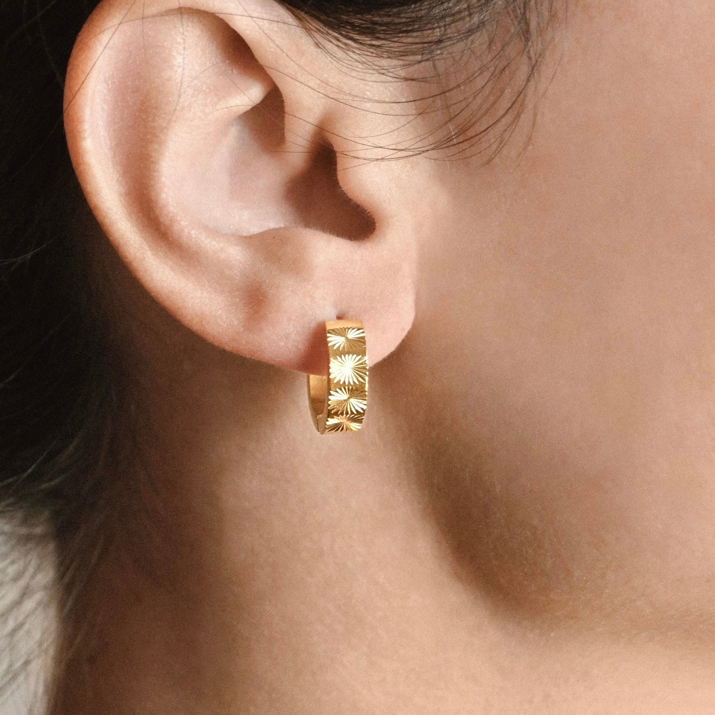 Close-up side profile of a woman with black hair, wearing small wide hoop earrings with starburst pattern on the  front.