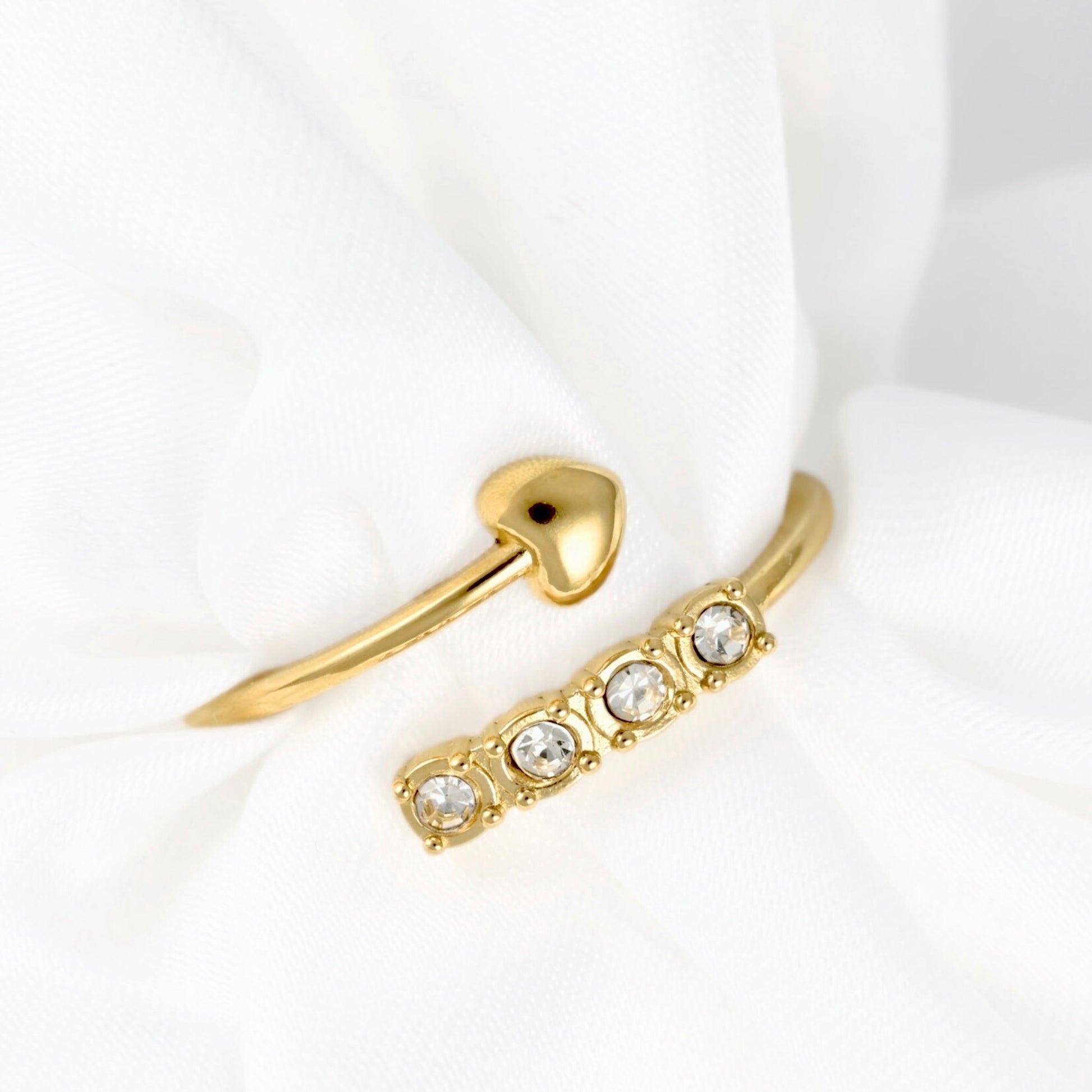A gold-toned ring set displayed on soft white fabric, featuring a slim band with three clear round gemstones and a matching small heart-shaped ring.