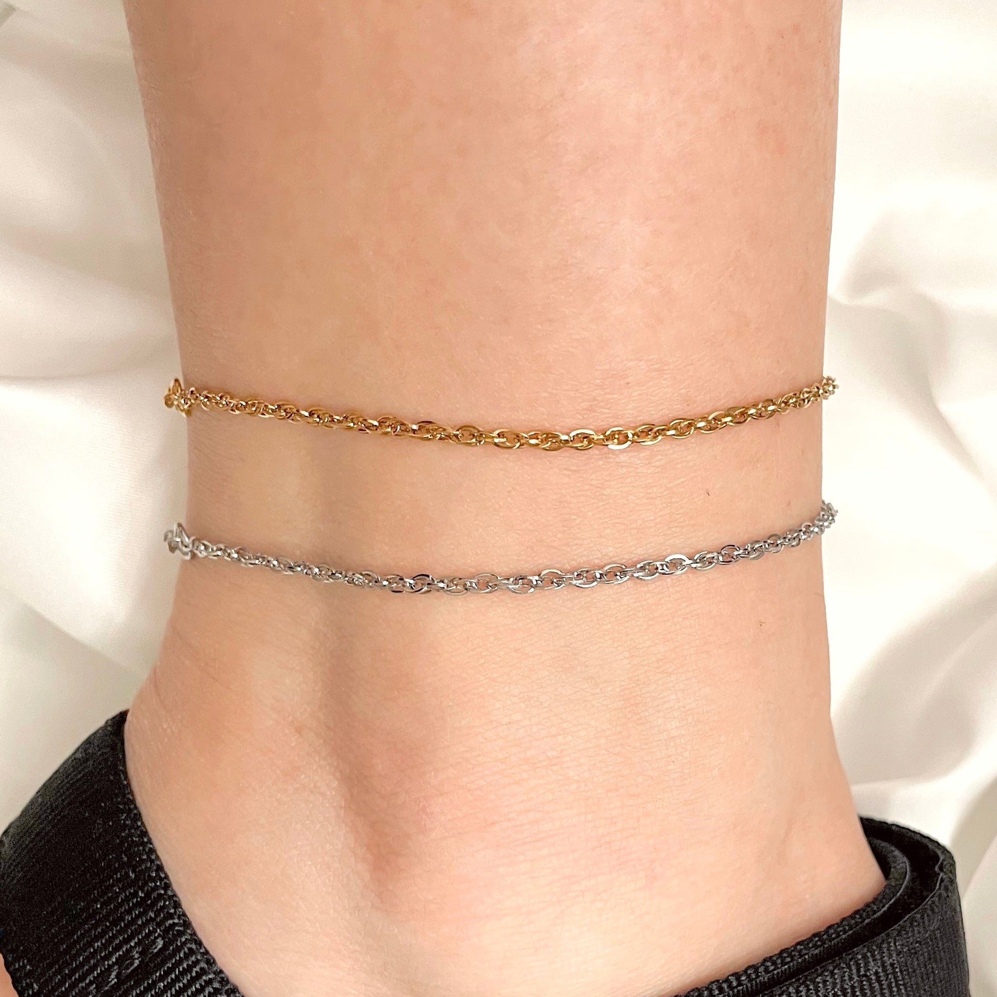 Two chain ankle bracelets, one gold and one silver, on a person's ankle against a white background.