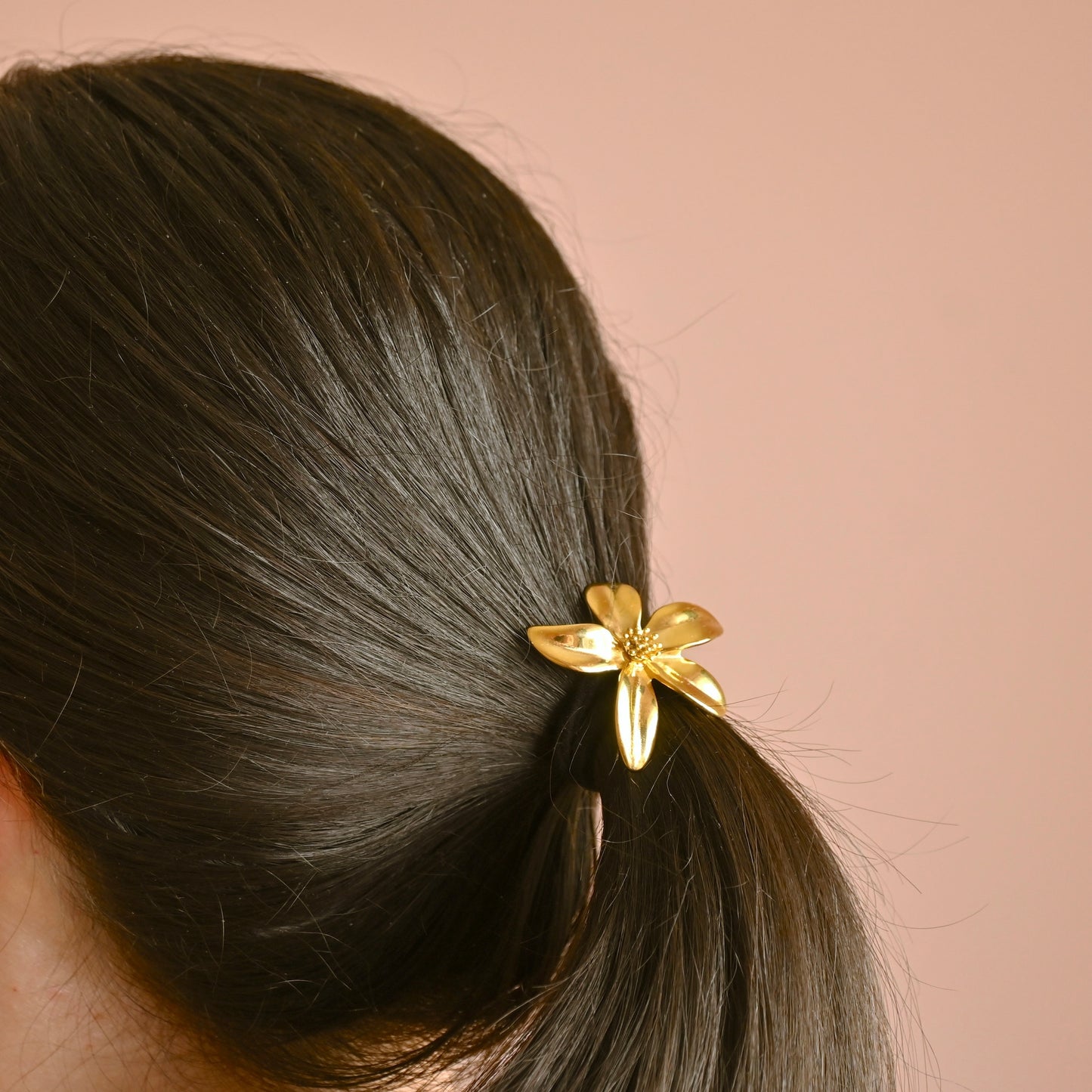 A close-up of a person's dark brown hair tied back in a low ponytail with a shiny gold flower-shaped hair tie, set against a solid dusty pink background.