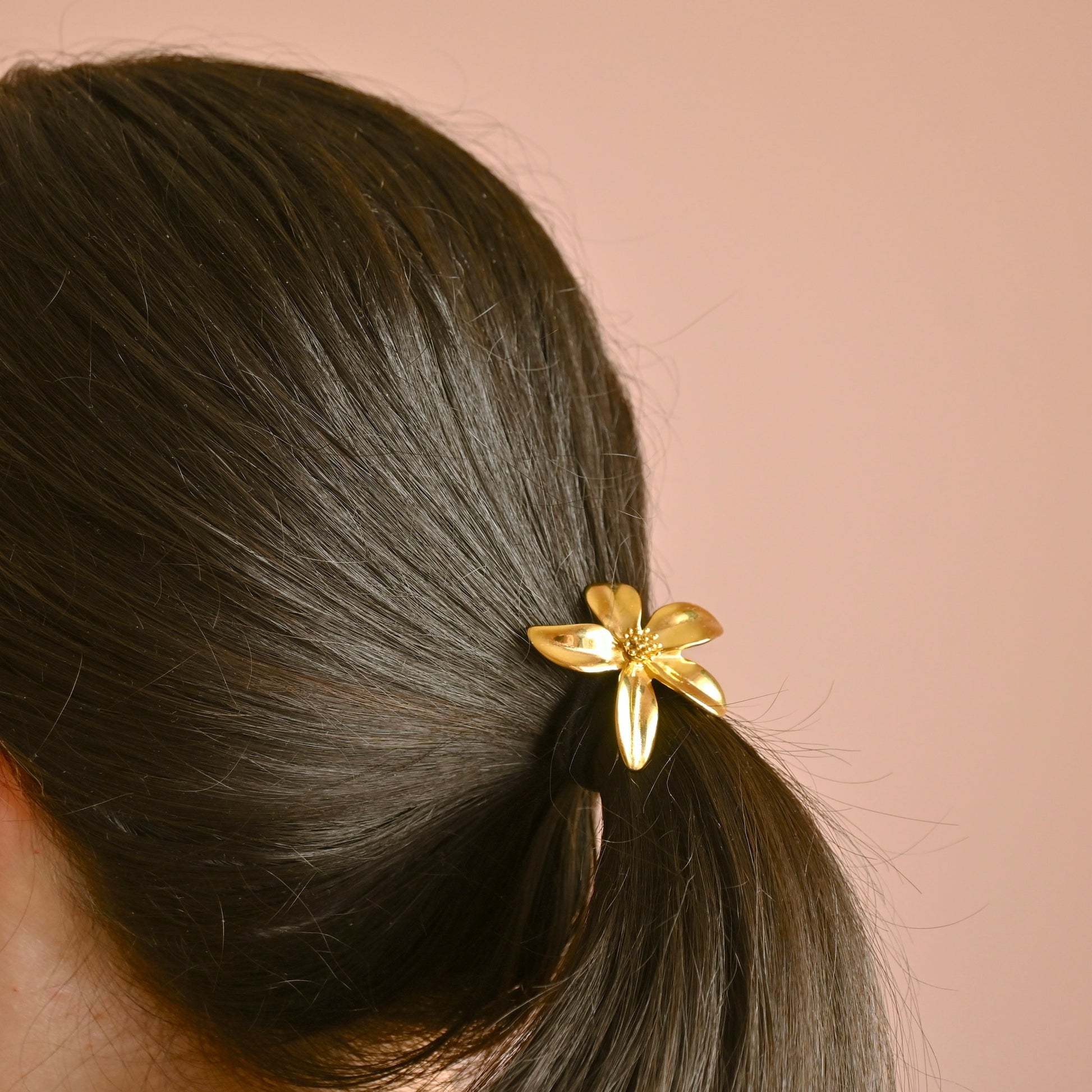 A close-up of a person's dark brown hair tied back in a low ponytail with a shiny gold flower-shaped hair tie, set against a solid dusty pink background.
