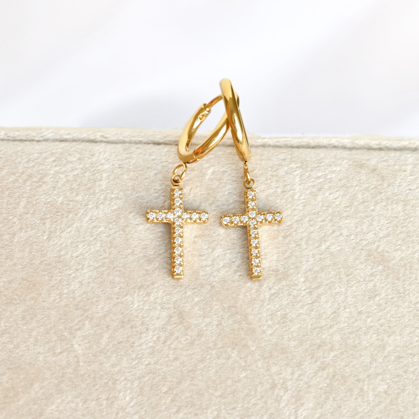 A pair of gold hoop earrings with dangling CZ cross charms, each adorned with small clear stones, displayed on a textured beige jewelry stand.
