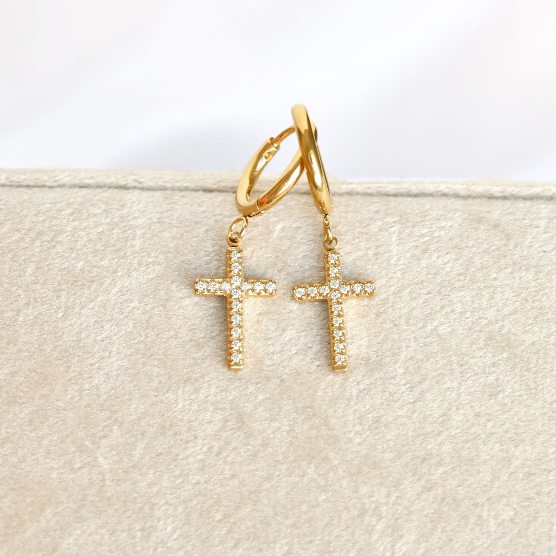 A pair of gold hoop earrings with dangling CZ cross charms, each adorned with small clear stones, displayed on a textured beige jewelry stand.