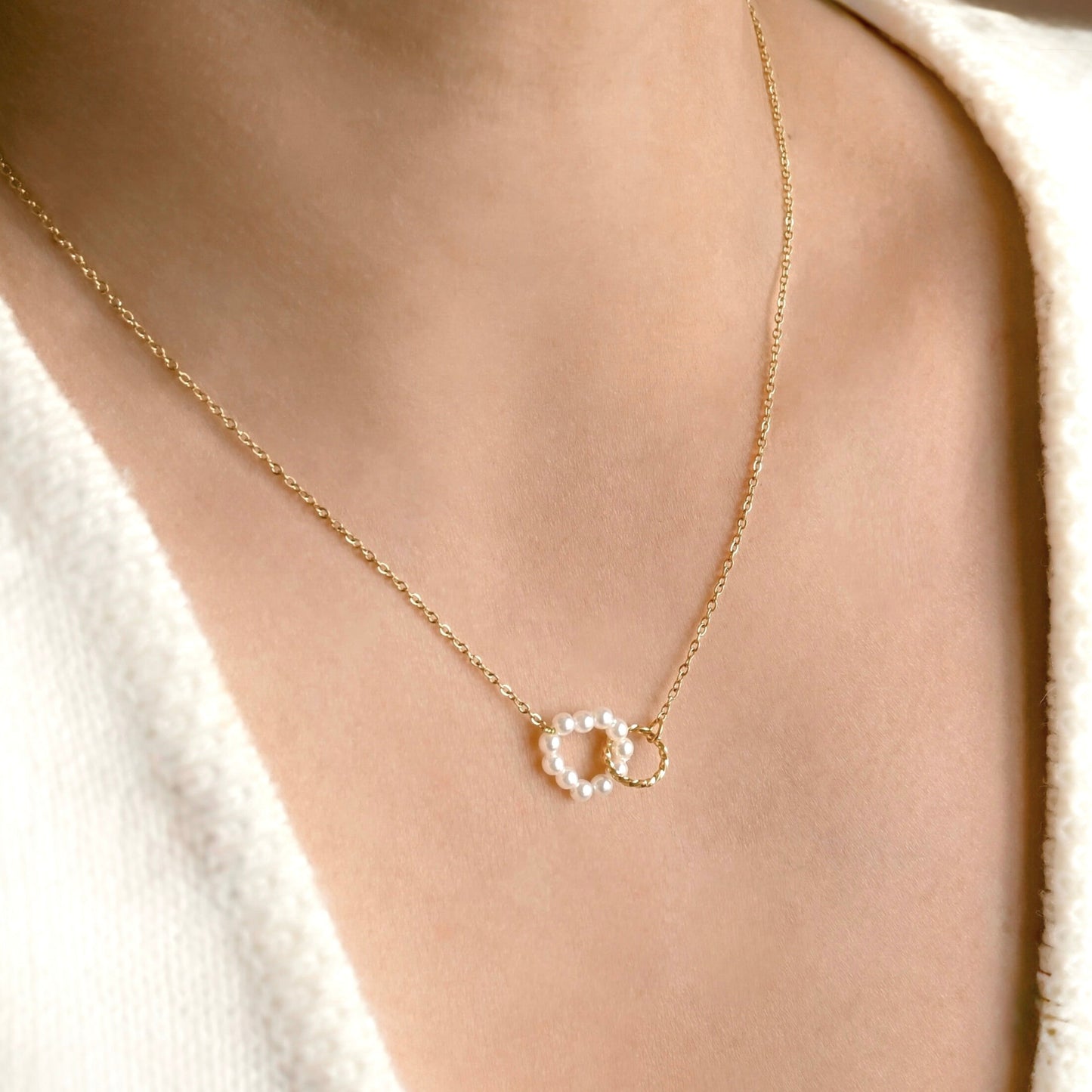 A close-up of a model wearing a delicate gold chain necklace with a double interlocking ring pendant, one of which is set with small clear crystals. The model is wearing a soft white knit top.