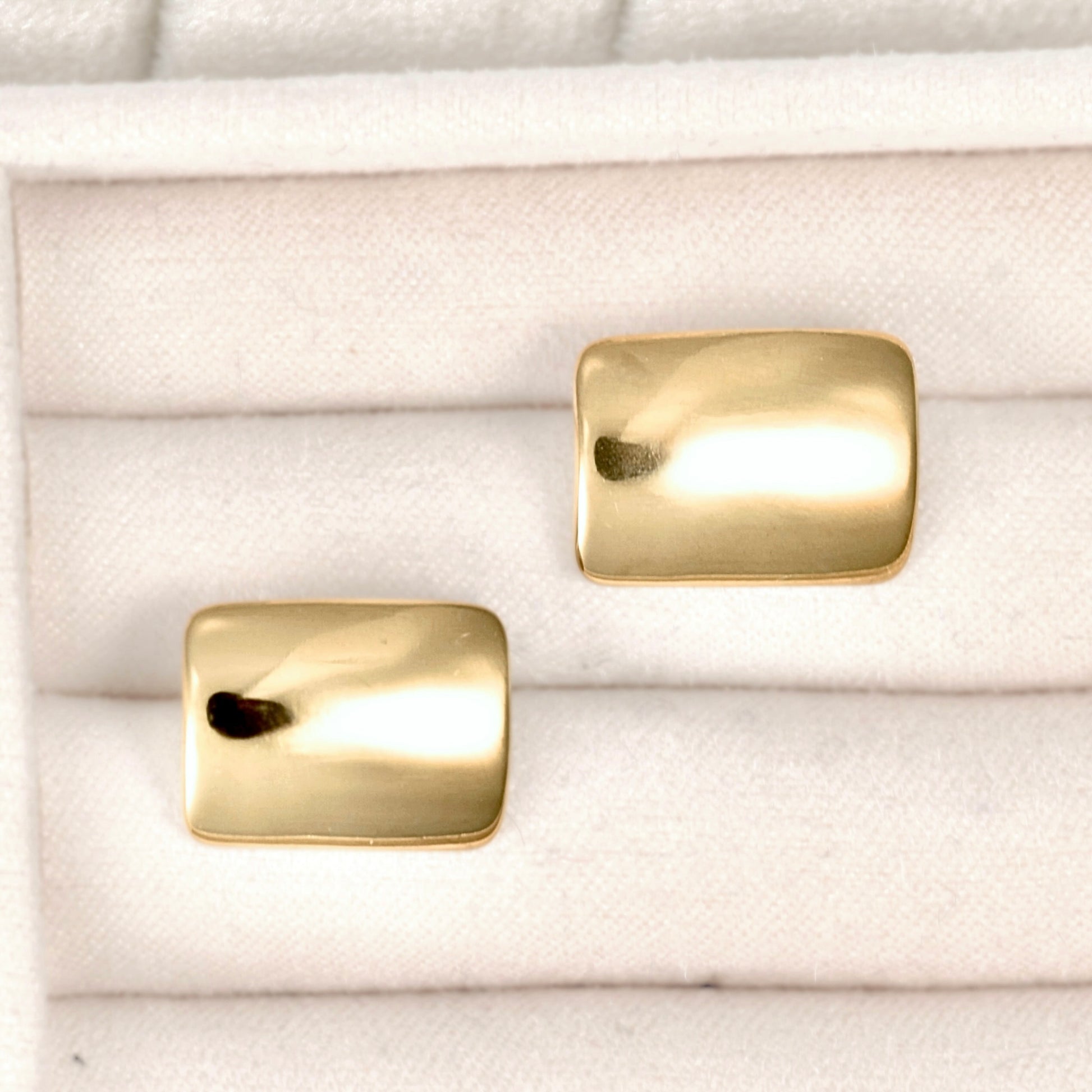 A pair of gold-toned rectangular stud earrings with a polished, slightly rounded finish, displayed on a white textured ring box.