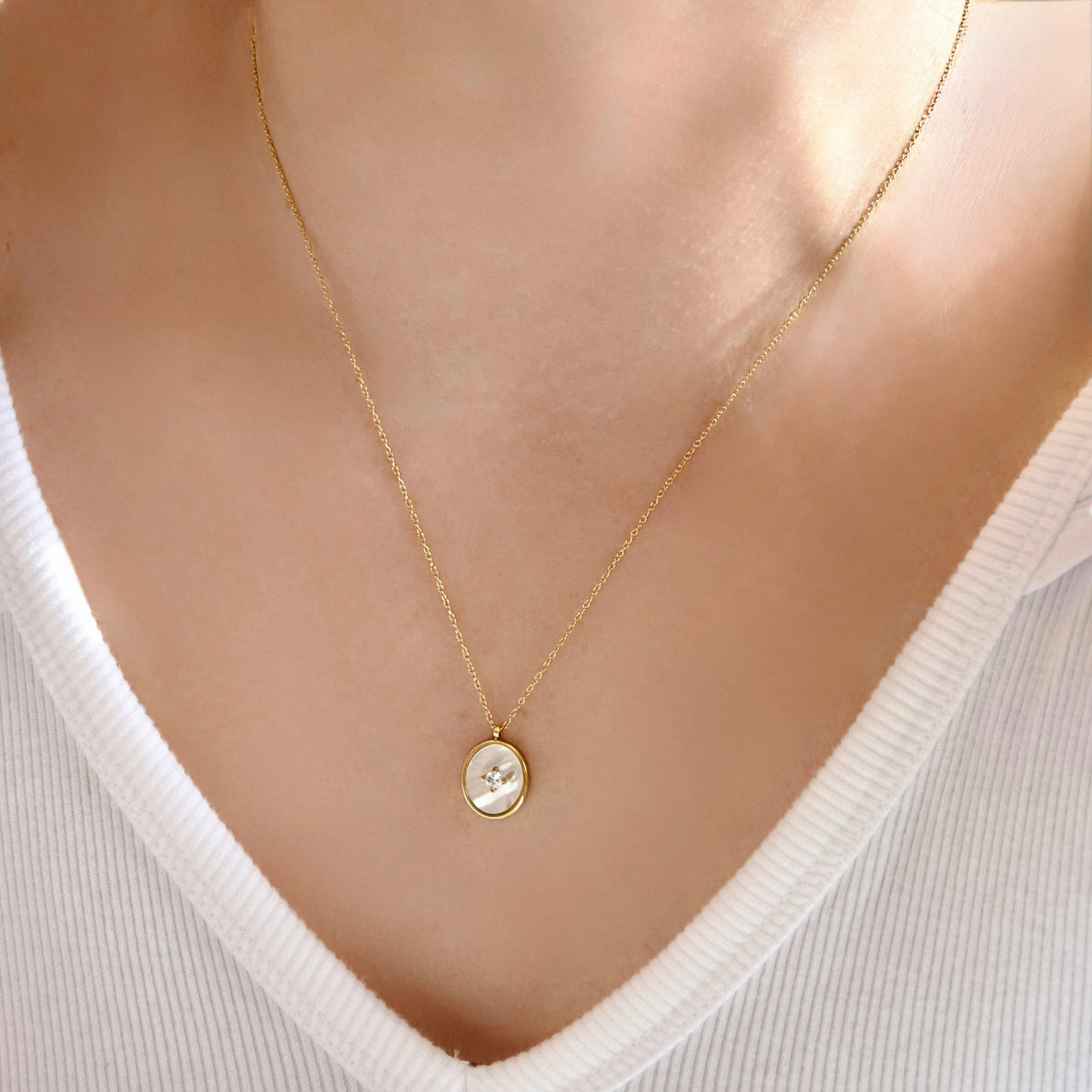 A delicate gold chain necklace with a small oval pendant featuring a white mother-of-pearl button, shown worn on a model's neck over a white ribbed V-neck top.