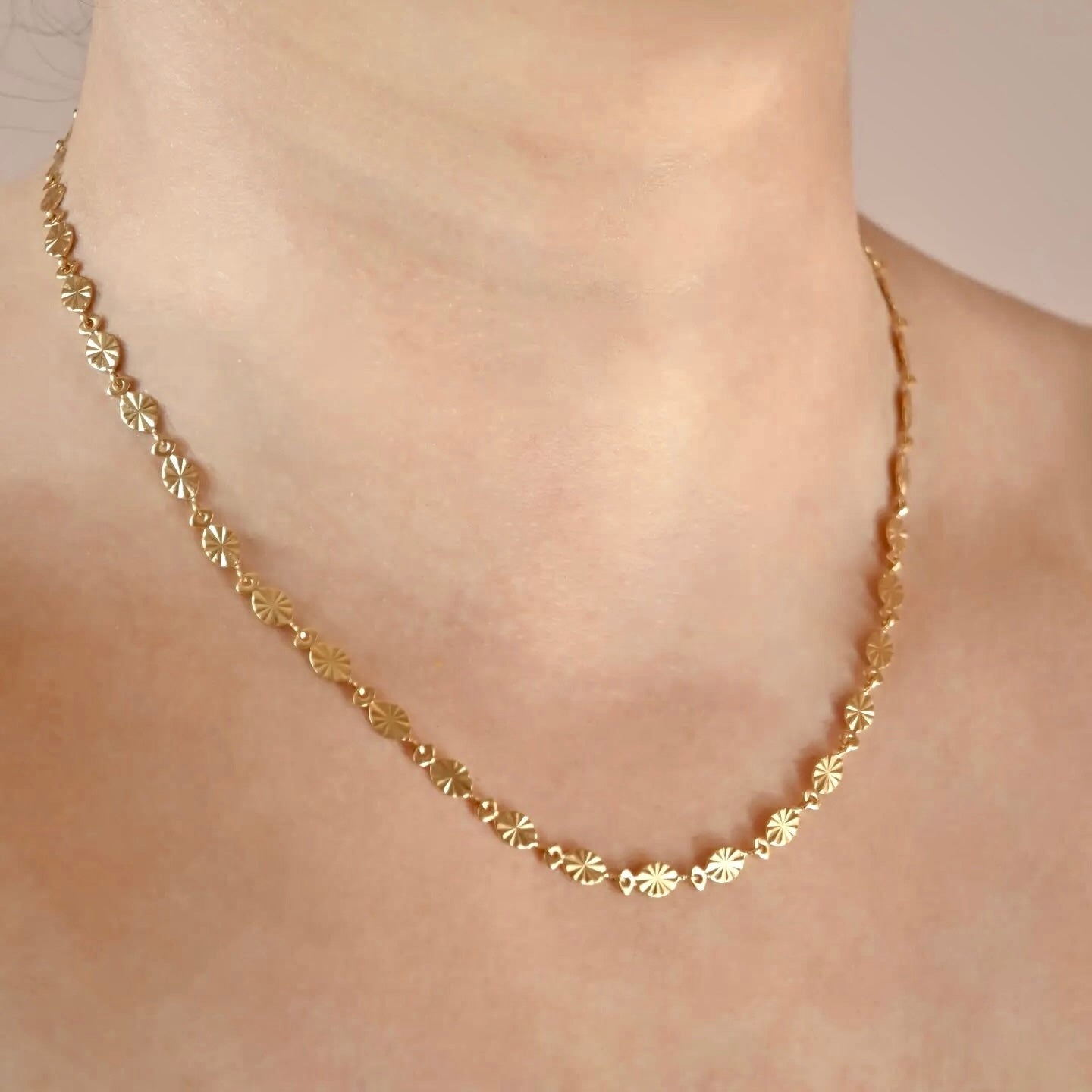 A delicate gold chain necklace with small, repeating sunburst motifs, shown worn on a person's neck and upper chest.