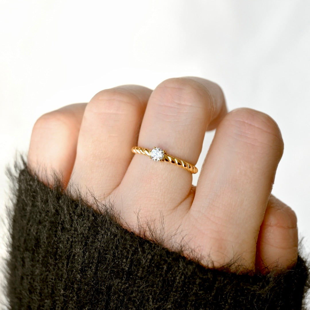 A close-up of a hand wearing a delicate twisted gold ring with a single round clear gemstone, set against a soft white background.