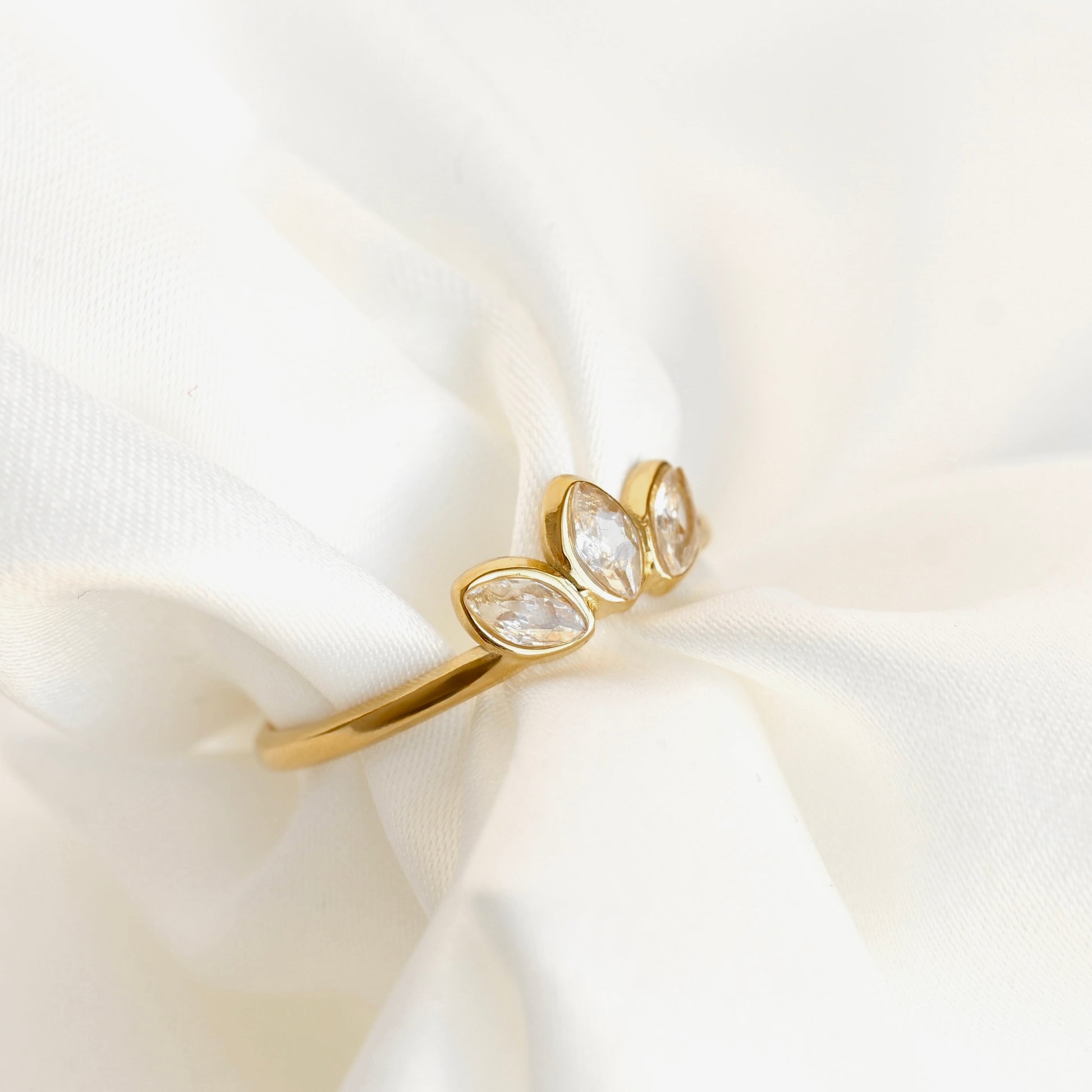 A gold ring with three marquise-cut clear gemstones set in a row, displayed on soft white draped fabric.