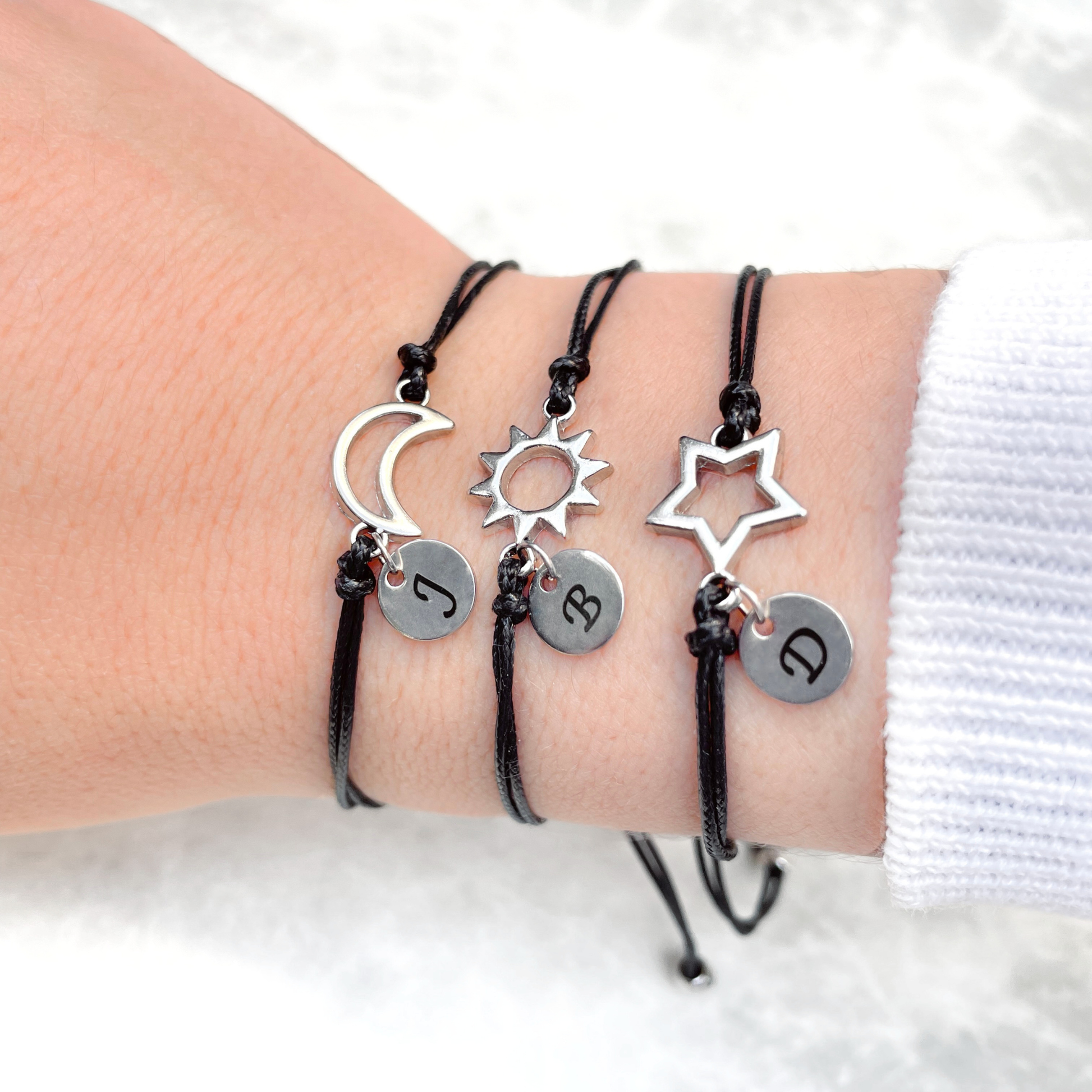 Three black cord bracelets with silver celestial charms and initials on a wrist against a white background