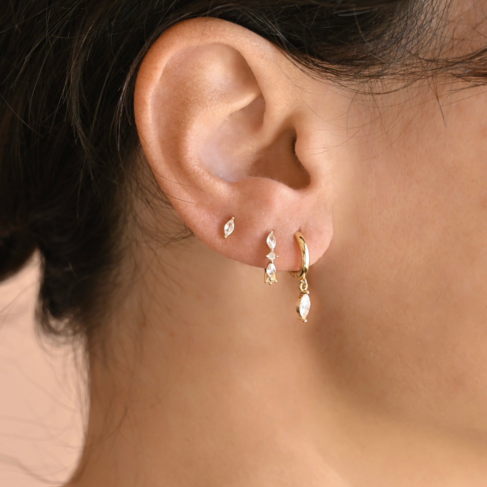 Close-up of an ear wearing three gold earrings, a drop charm hoop, huggie hoop and a stud with clear marquise stones.