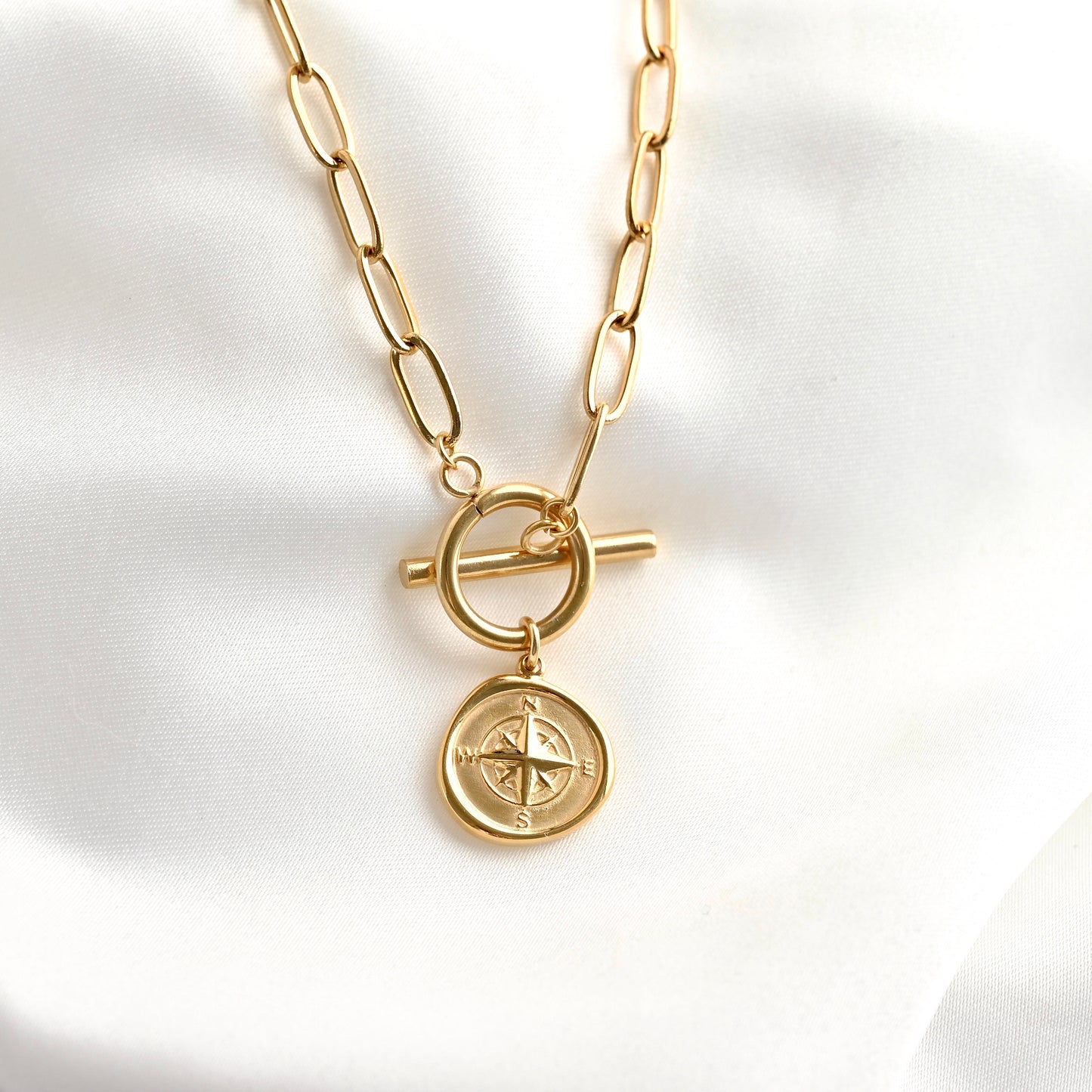 A gold-toned chain necklace with a toggle clasp and a round compass pendant, displayed on a soft white fabric background.