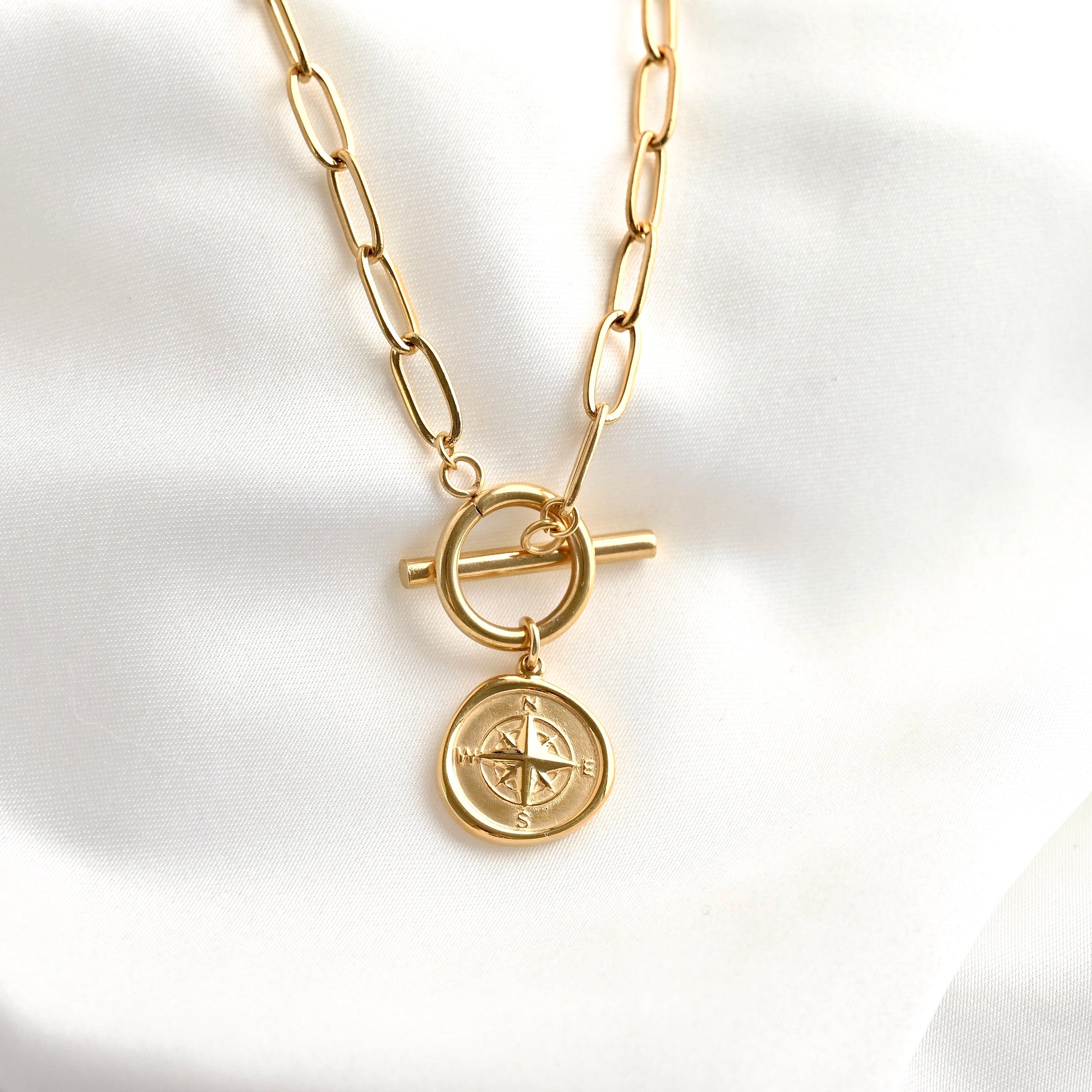 A gold-toned chain necklace with a toggle clasp and a round compass pendant, displayed on a soft white fabric background.
