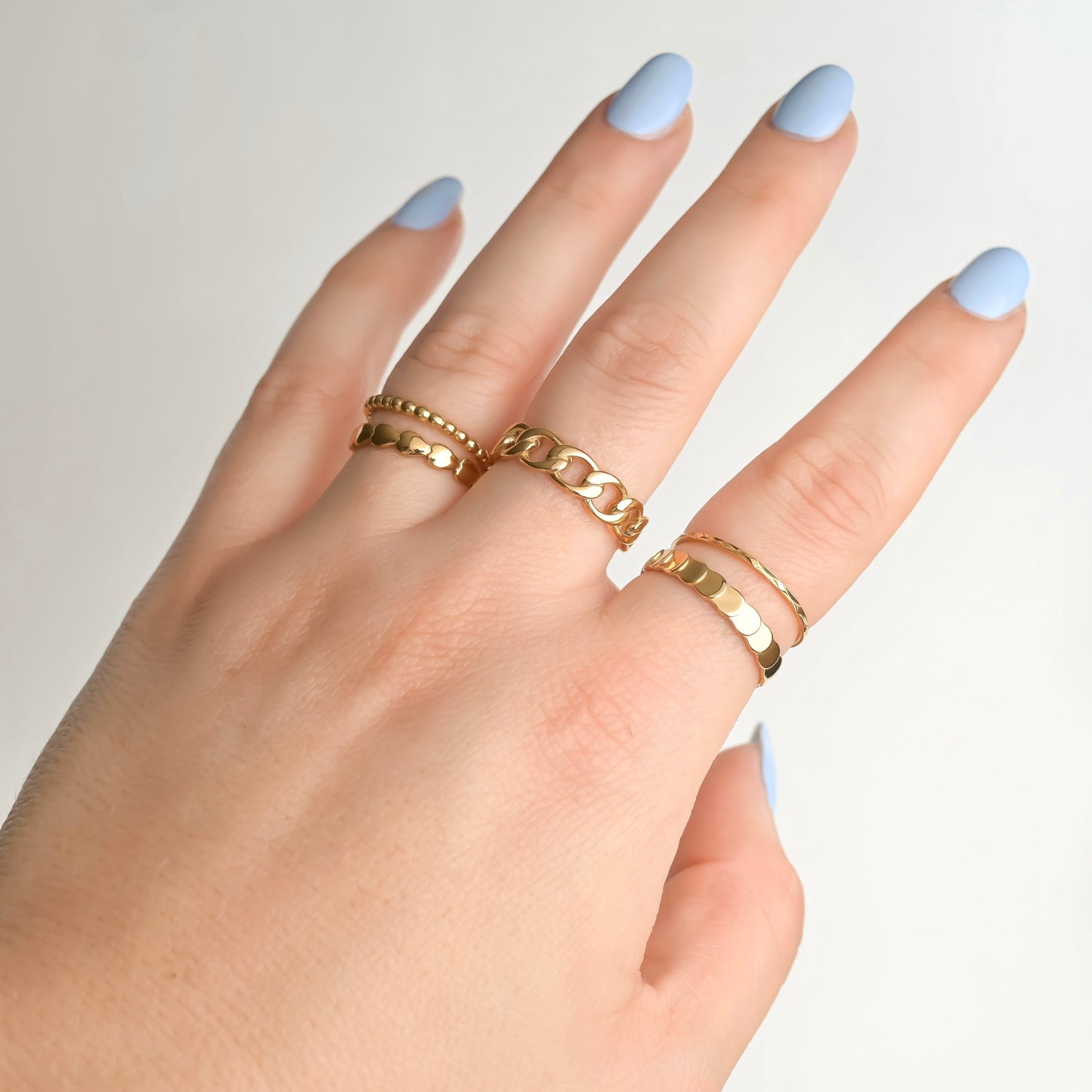 Hand wearing stacked gold rings with light blue nail polish on a neutral background