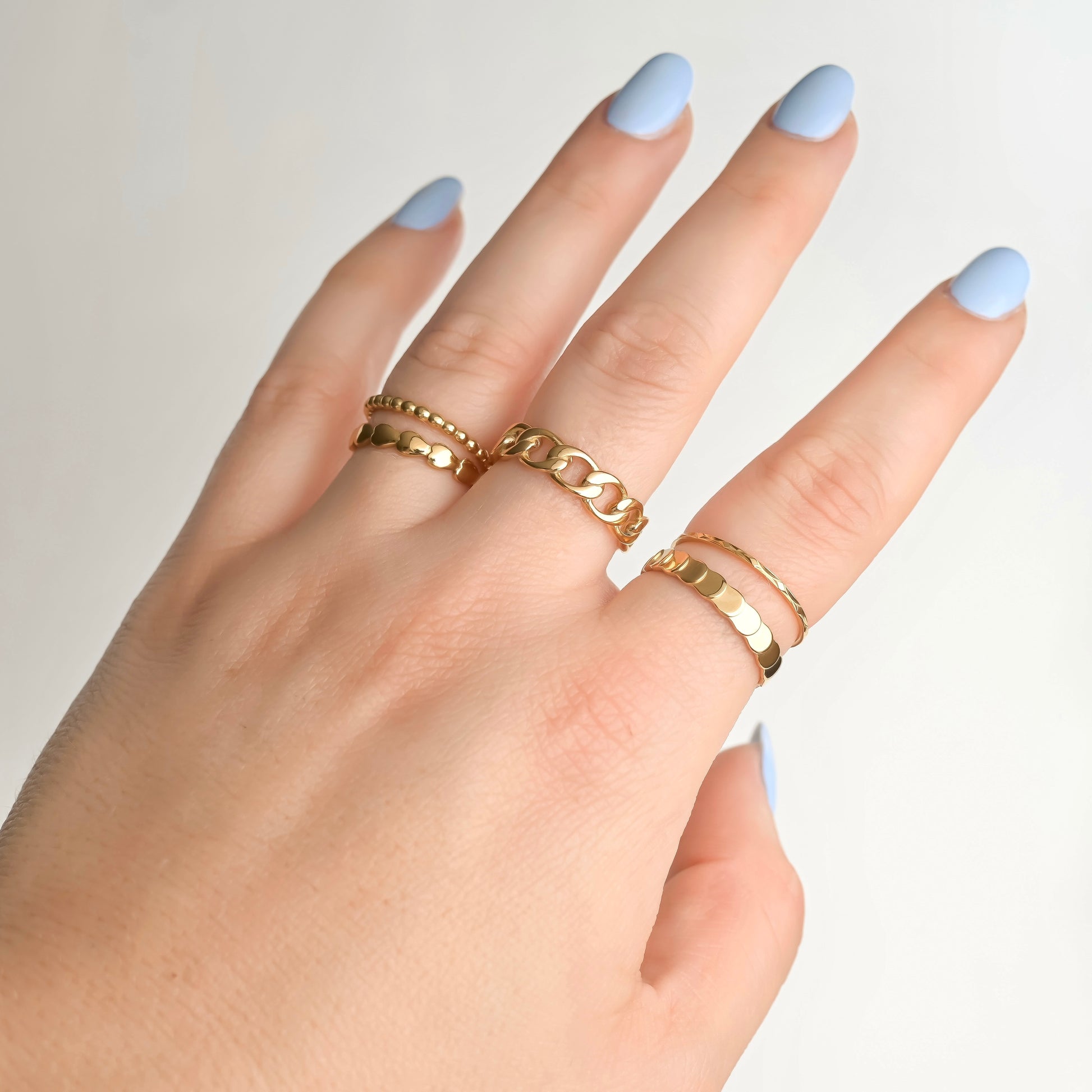Hand wearing stacked gold rings with light blue nail polish on a neutral background