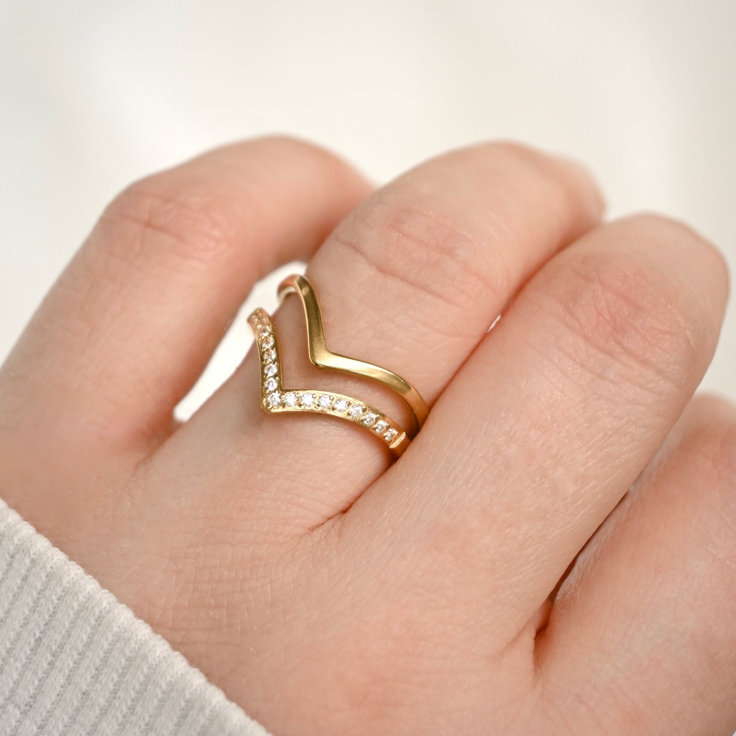 A gold ring with a double chevron design, one adorned with small pavé-set clear stones, worn on a person's finger.