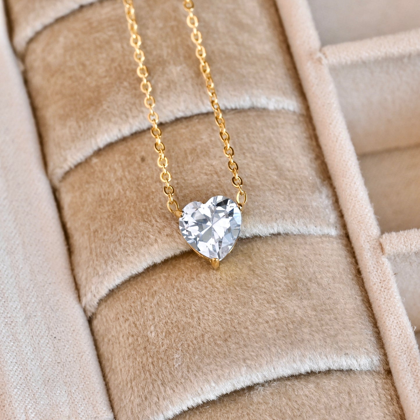 Gold necklace with a clear heart-cut  crystal pendant on a textured beige surface