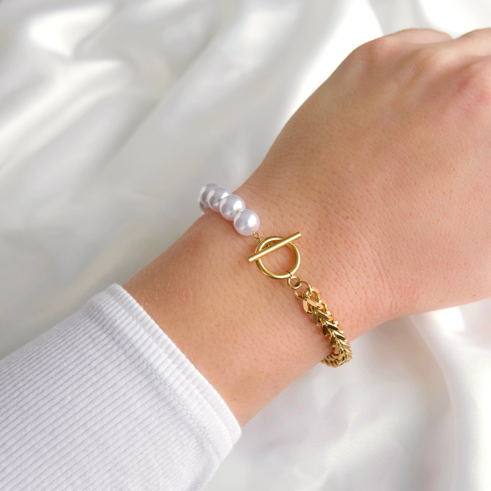 A close-up of a person's wrist wearing a gold-tone toggle clasp bracelet with a franco chain link and a strand of white faux pearls, set against a soft white fabric background.