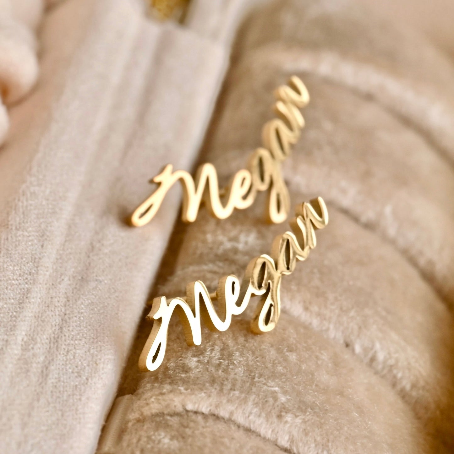 Gold 'Megan' name stud earrings on a beige fabric background