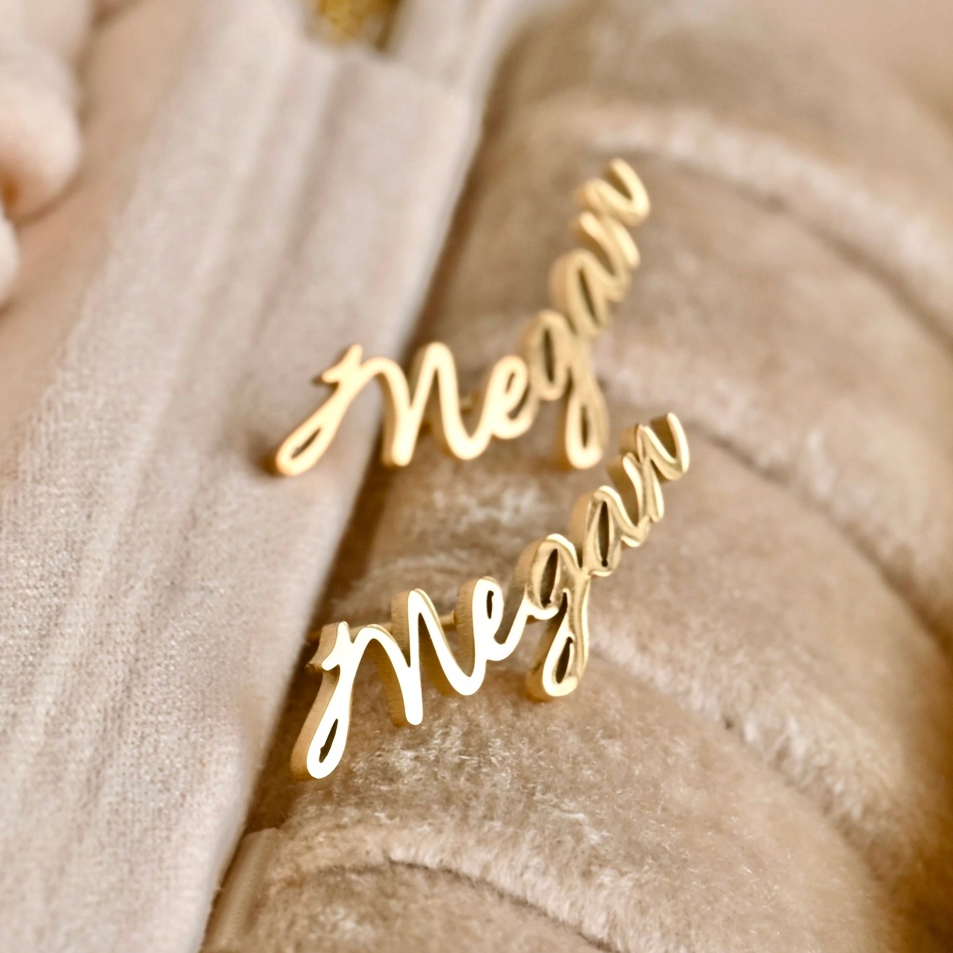 Gold 'Megan' name stud earrings on a beige fabric background