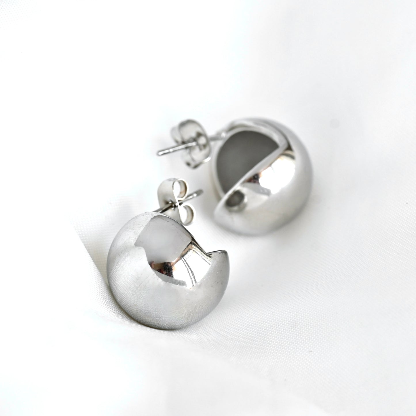 A pair of polished silver stud earrings shaped like stylized, open half-moon or crescent-shaped beads, displayed on a clean white fabric background.
