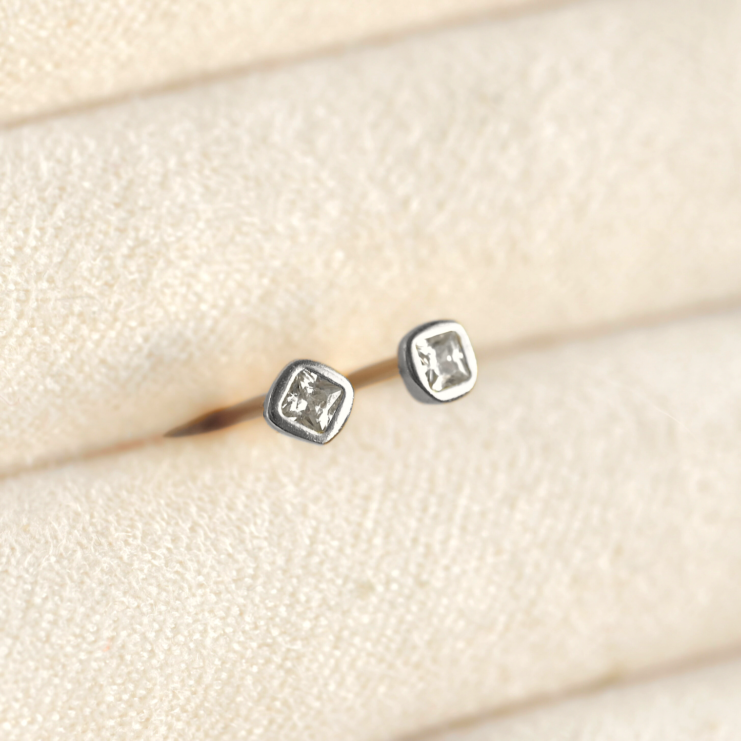 A pair of small silver stud earrings with square bezel-set clear CZ stones, displayed on a cream textured jewelry stand.