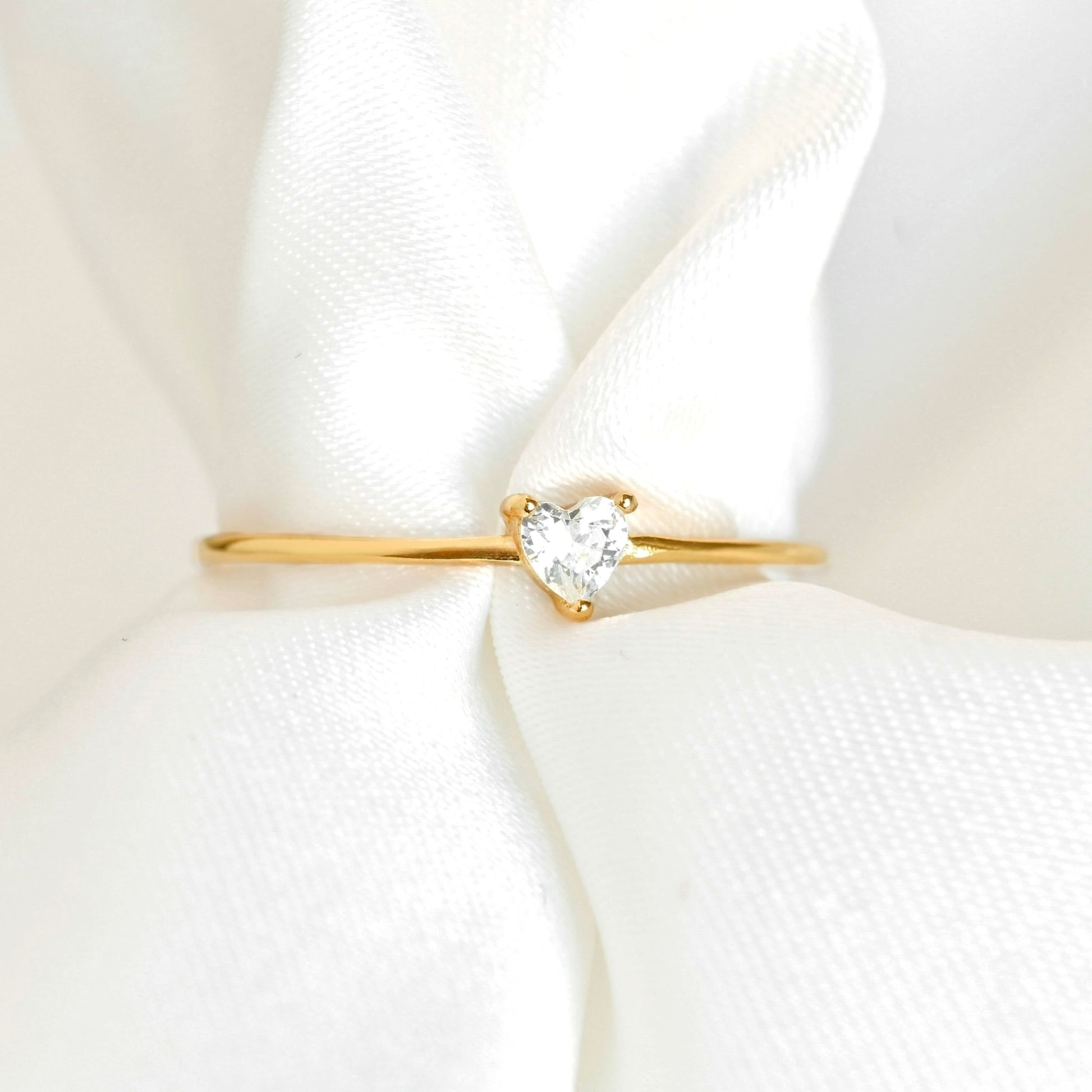 A dainty gold heart ring with a small heart-shaped clear gemstone, displayed on a textured white fabric background.