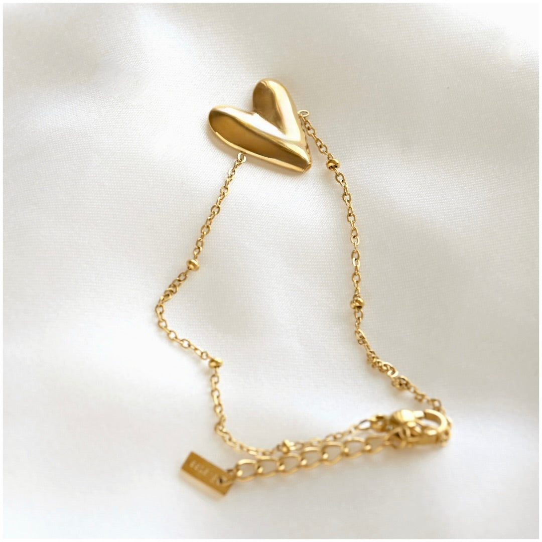 A gold chain bracelet featuring a big polished, three-dimensional heart charm at the top, displayed on a soft white fabric background.
