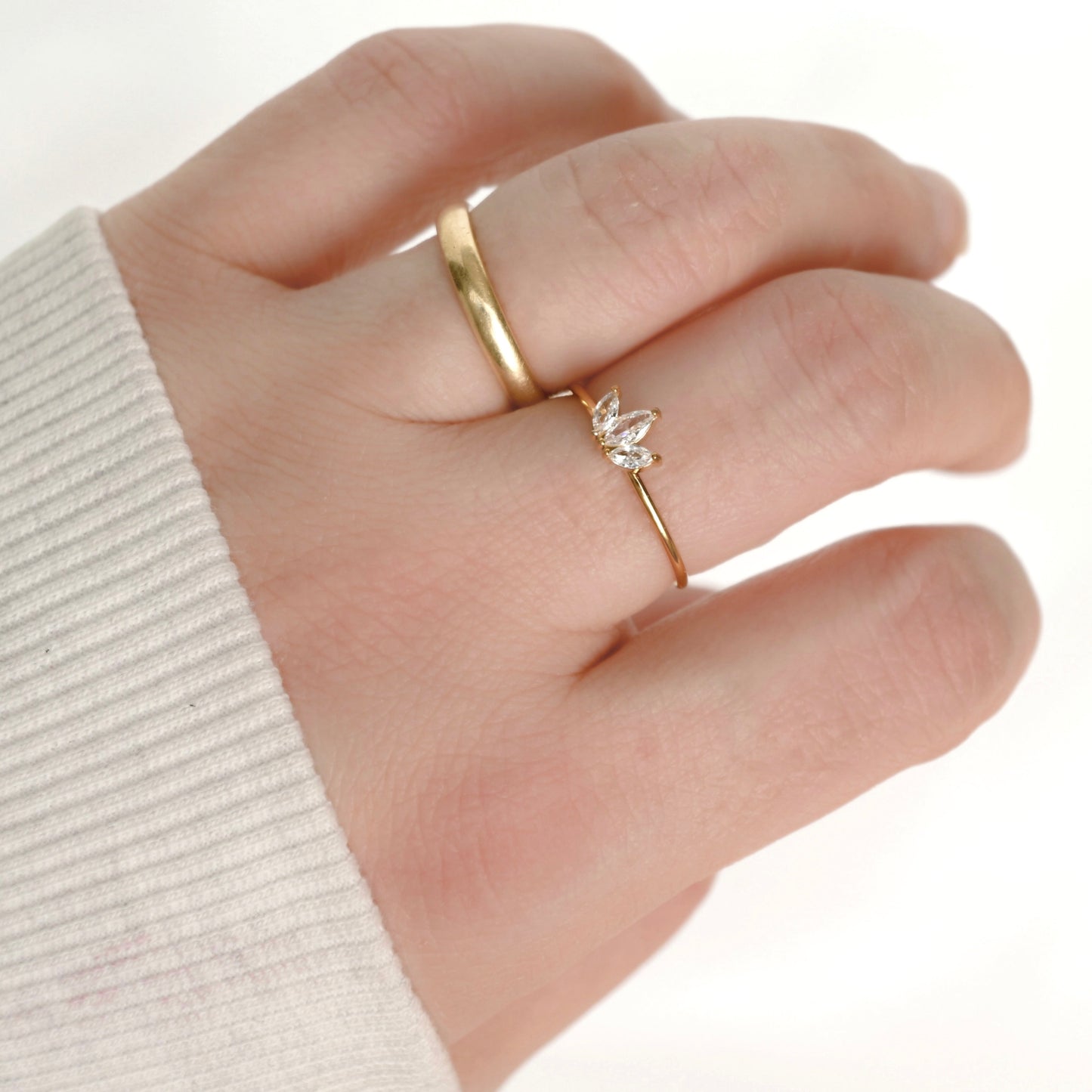 A close-up of a hand wearing two delicate gold rings, including a small ring with a clear crystal lotus flower setting, against a white background.