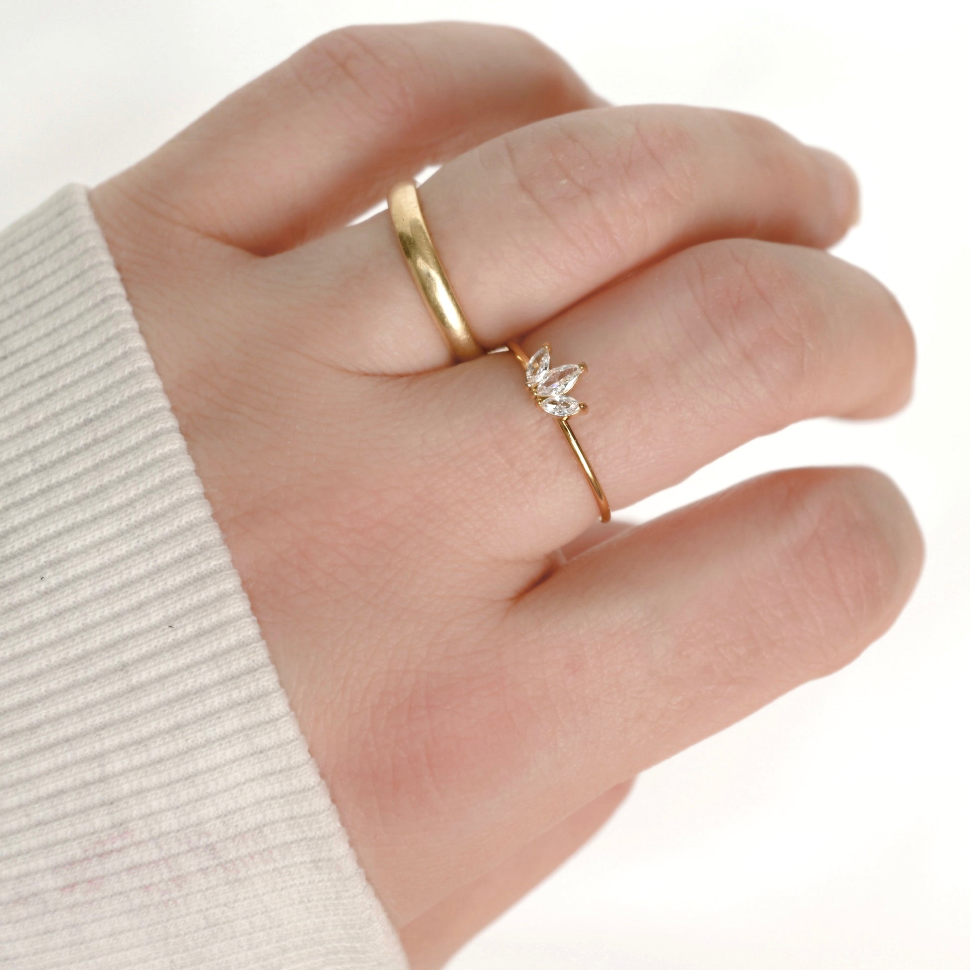 A close-up of a hand wearing two delicate gold rings, including a small ring with a clear crystal lotus flower setting, against a white background.