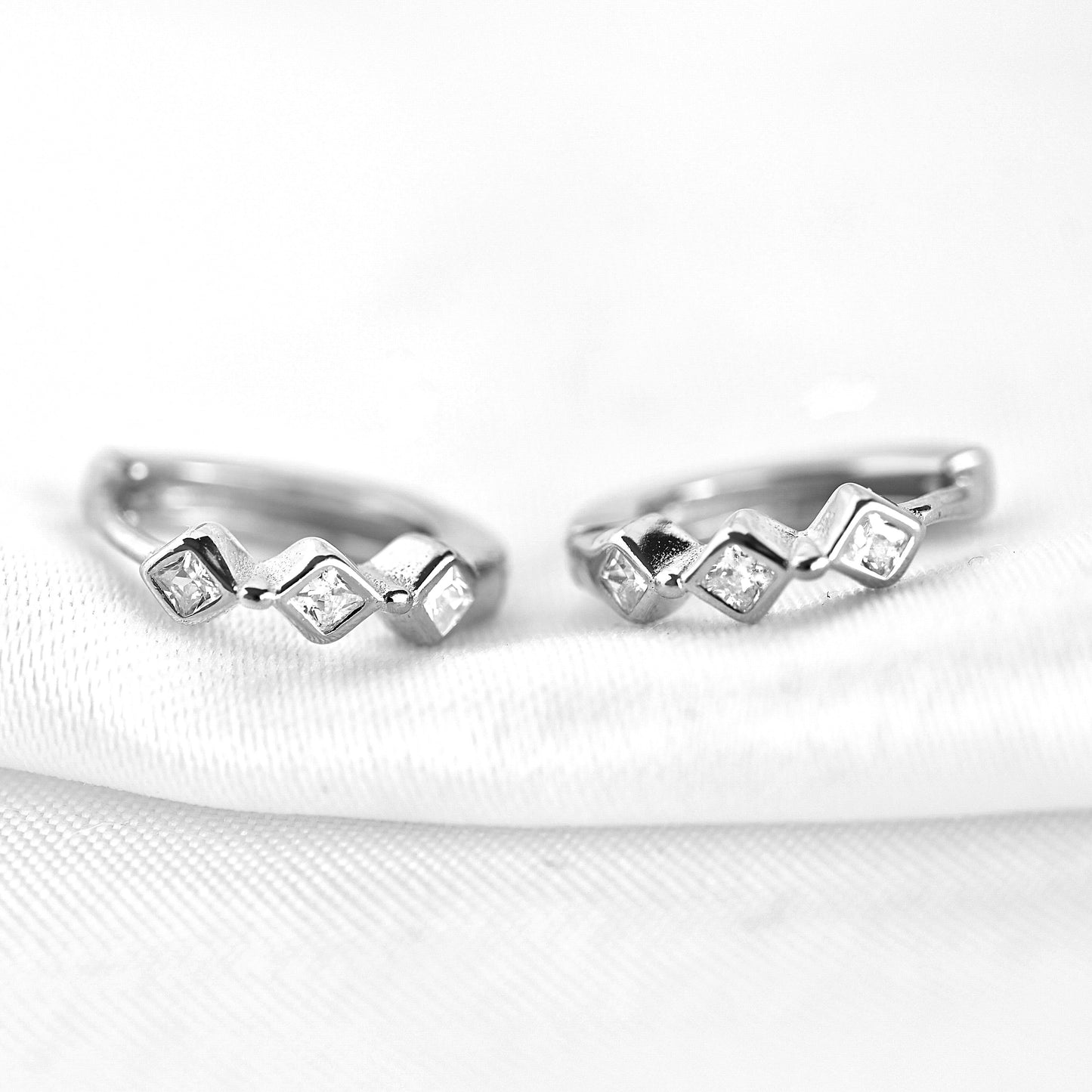 A pair of silver huggie hoop earrings with small clear gemstones set in a zigzag pattern, resting on a white textured fabric surface.