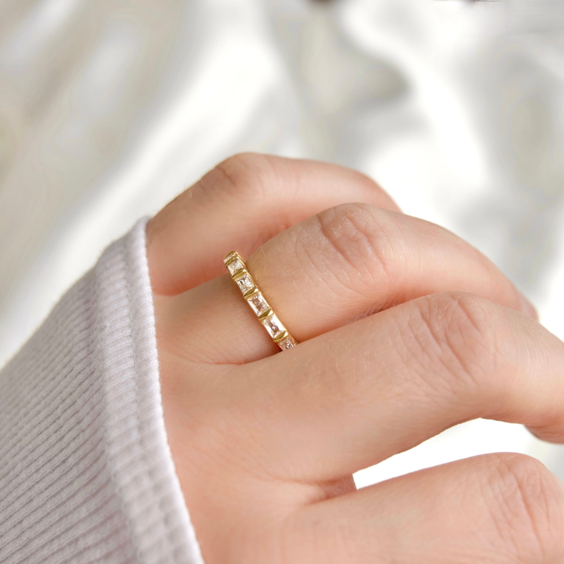A close-up of a hand wearing a delicate gold eternity ring set with small clear stones, styled with a white ribbed knit sleeve against a soft white fabric background.