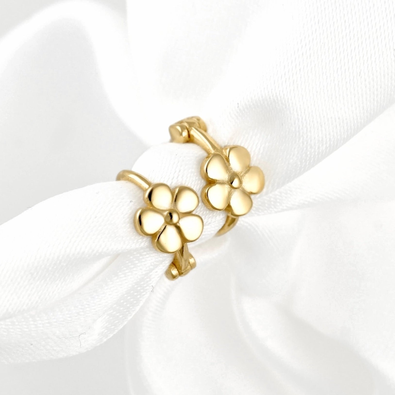 A pair of gold flower-shaped hoop earrings displayed on a soft white satin ribbon against a white background.